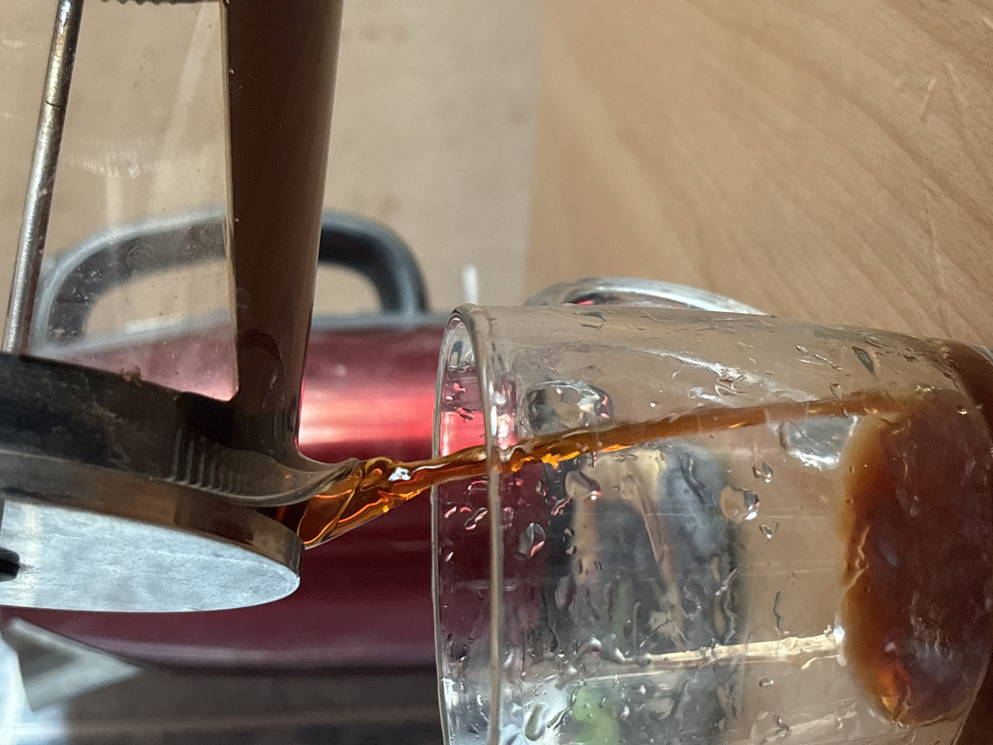 A close-up captures coffee being poured from a French press into a glass mug. The French press is on the left of the frame, while the mug is positioned to the right. The coffee stream is visible as it flows from the press into the mug. Water droplets cling to the exterior of the glass mug. The background is blurry, but hints of red appliance is visible. The scene is indoors, against a wooden surface. There is no information about the location in the city of Chaikovsky, Russia, in the image.