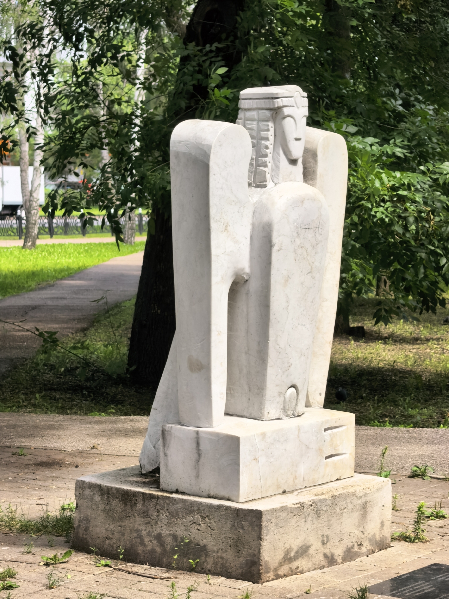 The image depicts a white stone sculpture of a stylized, possibly angelic figure with outstretched wings, located in a park-like setting. The sculpture is situated on a concrete base, which is itself placed on a paved area. Behind the sculpture, a curved concrete path is visible, bordered by lush green grass. Trees with dense foliage form a natural backdrop, with sunlight dappling through the leaves. In the distance, a fence and some trees are visible, suggesting a public area. The overall atmosphere is one of serene outdoor space. Based on the metadata, this sculpture is located in the city of Sterlitamak, Russia.