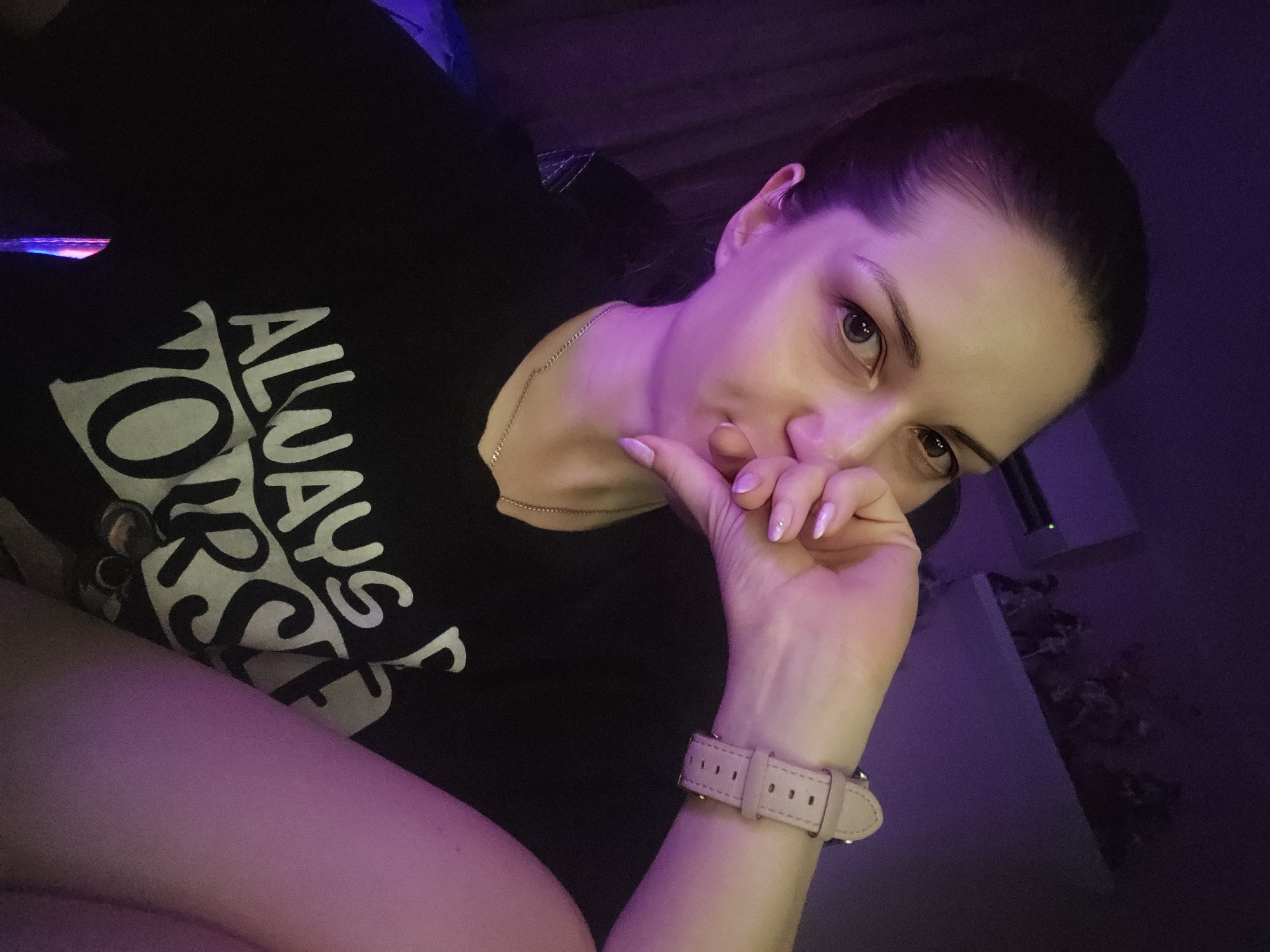 A woman with her hair pulled back is shown in a selfie, with her hand near her mouth. She is wearing a black t-shirt with white lettering that reads "ALWAYS YOURSELF". A silver necklace is visible around her neck, and she is wearing a light pink watch on her left wrist. The background is dark with purple lighting. The image does not provide any information about the city of Burgas, Bulgaria.