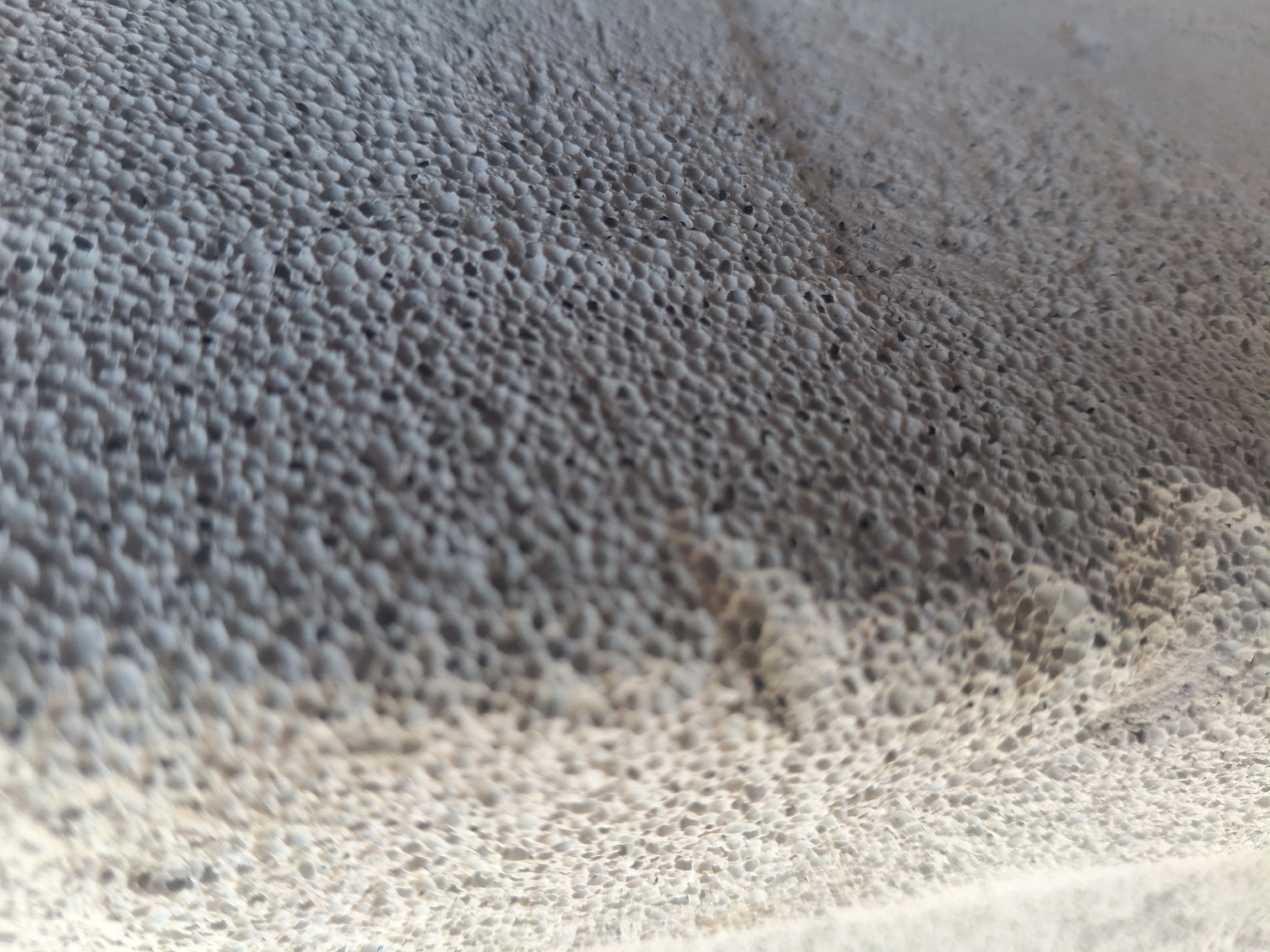 The image shows a close-up, textured surface of a light-colored material, possibly concrete or a similar building material, with a porous, pitted appearance. The texture is consistent across the visible area, with small, irregular indentations creating a honeycomb-like pattern. The lighting suggests an outdoor environment, casting subtle shadows within the pits and highlighting the raised surfaces. There are no people, animals, or specific landmarks visible. The overall impression is one of an abstract detail of an urban surface. The location in Kalush, Ukraine, is not identifiable from the visual information provided in the image.