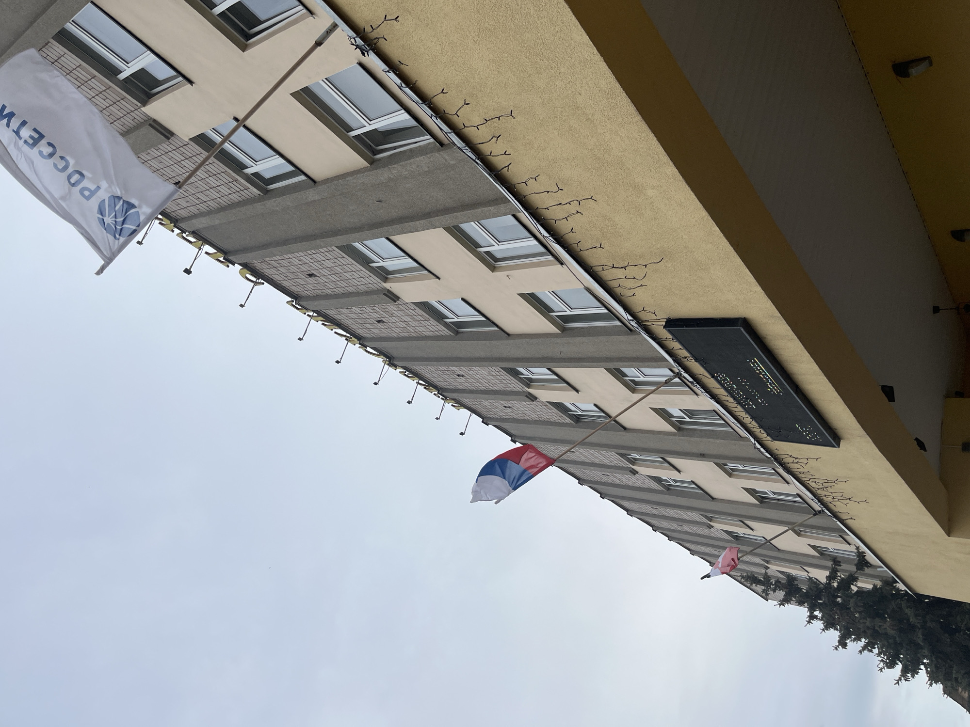 The image shows an upward view of a multi-story building in Izhevsk, Russia. There are three flags hanging from the building. The first flag on the left is white with a blue logo and the text "РОССЕТИ" (Rosseti). The second flag is the Russian tricolor flag of white, blue, and red. The third flag also appears to be the Russian tricolor flag. A black sign is visible attached to the building. The building is a beige color with gray accents. The sky is overcast and gray. The top right corner of the image includes the top of the building. Some tree branches are visible in the bottom right corner.