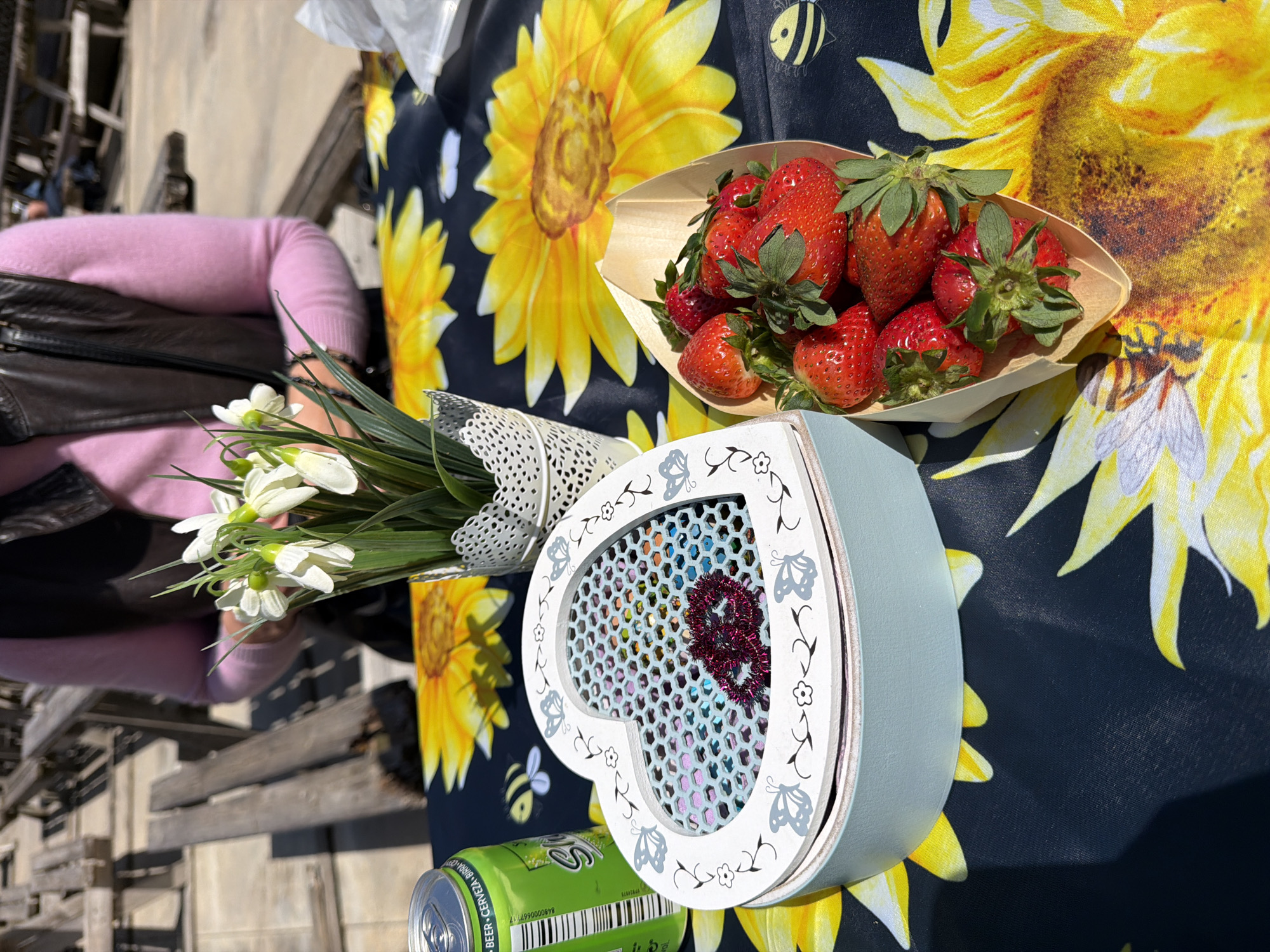 This image presents a vibrant outdoor scene, likely in Monistrol de Montserrat, Spain, featuring a collection of items on a table covered with a striking dark tablecloth patterned with large yellow sunflowers and small bees. The bright sunlight suggests it's daytime.
In the center, a small wooden boat-shaped container is filled with fresh, ripe red strawberries, their green caps still attached. Adjacent to this, a light blue heart-shaped box with a white, decorative lid captures attention. The lid is adorned with delicate cut-out designs of butterflies and small flowers, and features a grid-like opening revealing colorful foil squares and a sparkly red number '8' inside.
To the left, a bouquet of white flowers, possibly snowdrops or similar spring blossoms, is artfully wrapped in a white, lace-patterned material. A person is partially visible behind these flowers and to the left, wearing a light pink long-sleeved top and a dark vest or jacket. Their arm is near the bouquet.
A green beverage can is partially visible in the bottom left corner. Text on the can clearly reads "BEER", "CERVEZA", "BIRRA", and "DAMM", along with a barcode. The background shows blurred wooden structures, likely benches or railings, indicating a public outdoor setting. The overall impression is cheerful and pleasant, possibly a picnic or a market stall display.