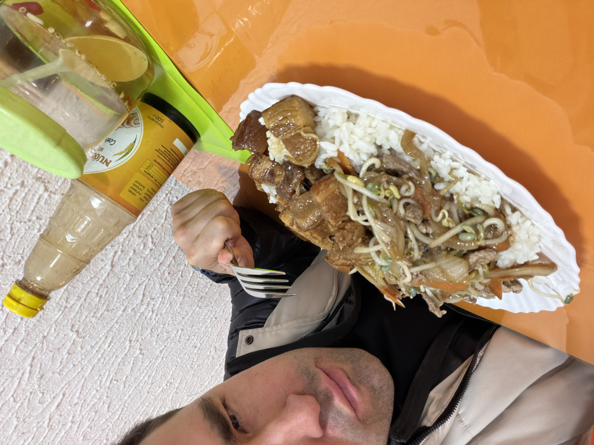 A man is depicted from a top-down perspective, positioned to eat a meal in an indoor setting. His face, with short dark hair and stubble, is visible in the lower right of the frame, showing a neutral expression. He is wearing a dark jacket with a lighter-colored inner layer. His right hand holds a metal fork, ready to scoop food from a white, scalloped-edge plate.
The plate holds a generous serving of white rice next to a stir-fry or stew. This dish features substantial chunks of meat, possibly pork belly, accompanied by stir-fried vegetables such as bean sprouts, sliced carrots, and other green components, all coated in a glossy, dark sauce.
To the left, a bright green plastic tray or placemat supports a mostly empty clear plastic bottle with a yellow cap and a yellow-labeled plastic container. The label on the container is partially visible, showing the brand "NƯỚC" and "Coel". Critical Russian text includes "Масса нетто 500 г" (Net weight 500g) and ingredients like "соль, сахар, колер, глутамат натрия, кислота, бензоат" (salt, sugar, colorant, monosodium glutamate, acid, benzoate), indicating a food product, possibly a sauce or condiment, commonly found in Krasnoyarsk, Russia.
The immediate surroundings feature a textured, light-colored surface (likely a wall or patterned tablecloth) on the left, and a smooth, bright orange tabletop on the right, upon which the food plate and parts of the green tray are placed. The scene conveys a casual, everyday meal.