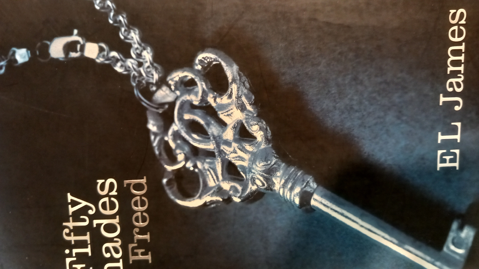 The image is a close-up shot of the cover of the book "Fifty Shades Freed" by E.L. James. The dominant visual element is a metallic key pendant on a silver chain, positioned diagonally across the frame. The key has an ornate, decorative head with intricate loops and detailing. The chain is made of interlocking links, with a clasp visible at the top right of the image. The background is a dark, textured surface, possibly a book cover itself, with a subtle gradient of dark blue and black tones. The title "Fifty Shades Freed" is printed in white, stylized lettering on the upper left side of the cover, with "Freed" being the largest of the words. Below the key and chain, also in white lettering, is the author's name, "EL James". There are no people or any indication of a location in the city of Kos, Greece.