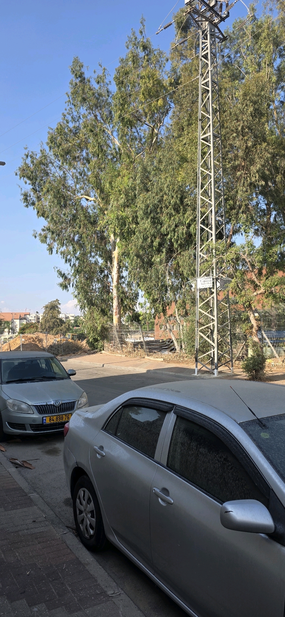 In Sderot, Israel, two silver cars are parked on the side of a paved street, with one vehicle in the foreground and another slightly behind and to the left. A tall, industrial metal pylon with electrical insulators and wires stands prominently to the right of the frame, casting a shadow on the street. Large trees with green foliage surround the area, with a construction site visible in the background to the left, featuring mounds of dirt and a yellow bulldozer. The sky is clear and blue, indicating a daytime scene.