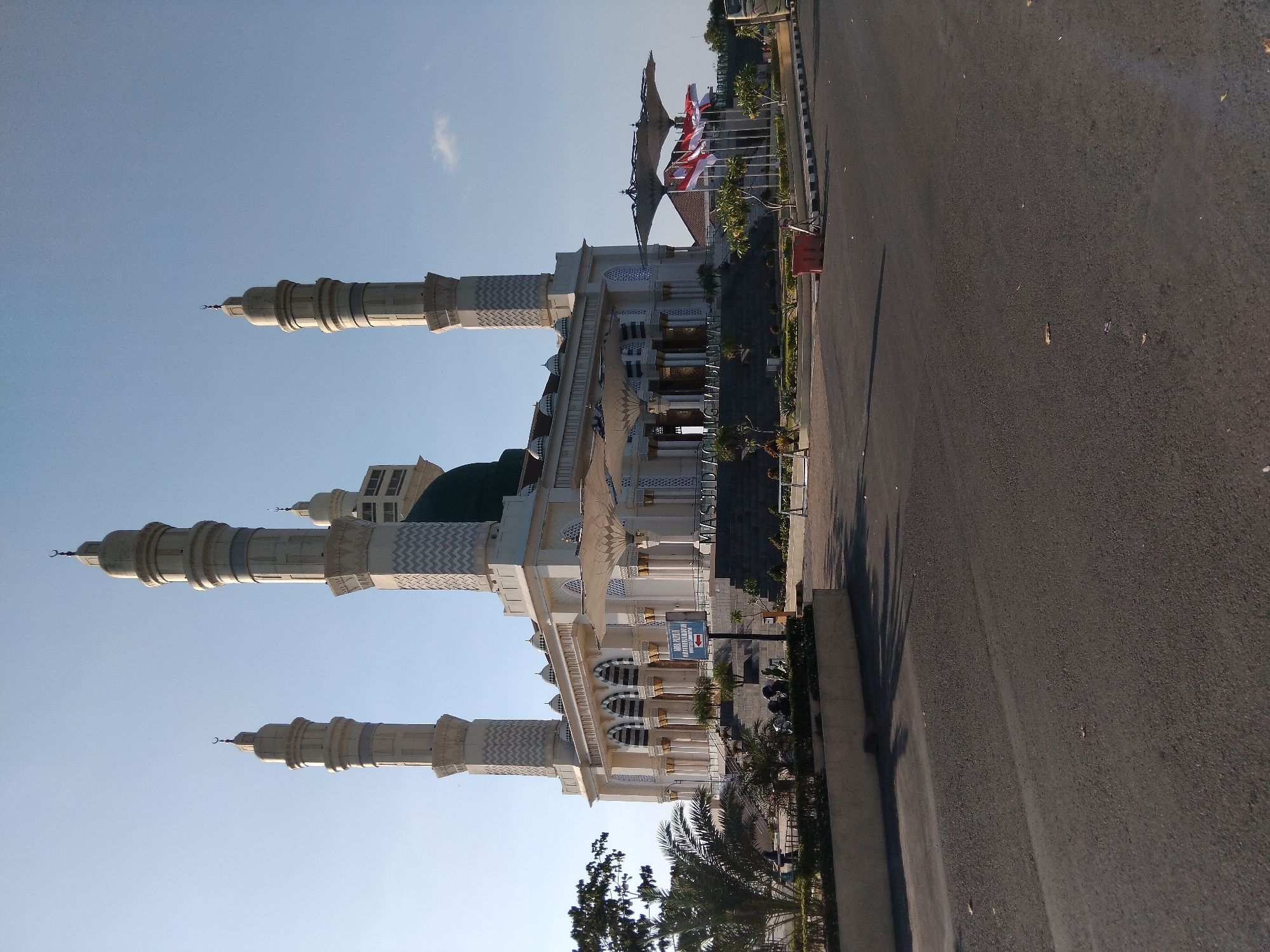 The image captures a wide shot of the Grand Mosque of Madinatunnur, located in Karanganyar, Indonesia, under a clear, bright sky. The mosque is a prominent structure featuring a large green dome and four tall, slender minarets that dominate the skyline. The building itself is primarily white with intricate architectural details, including arched windows and decorative patterns on the minarets. In front of the mosque, several white, umbrella-like canopies provide shade.
To the right of the mosque, a row of flags, including the Indonesian flag (red and white), are prominently displayed. In the foreground, a paved road stretches across the image, with a landscaped area featuring trees and planters between the road and the mosque complex. A signpost with an arrow pointing to the left is visible near the entrance to the mosque. A few individuals are seen in the distance, walking near the mosque's entrance, but they are not the primary focus of the image. The overall scene conveys a sense of grandeur and religious significance, set against a serene, sunny backdrop.