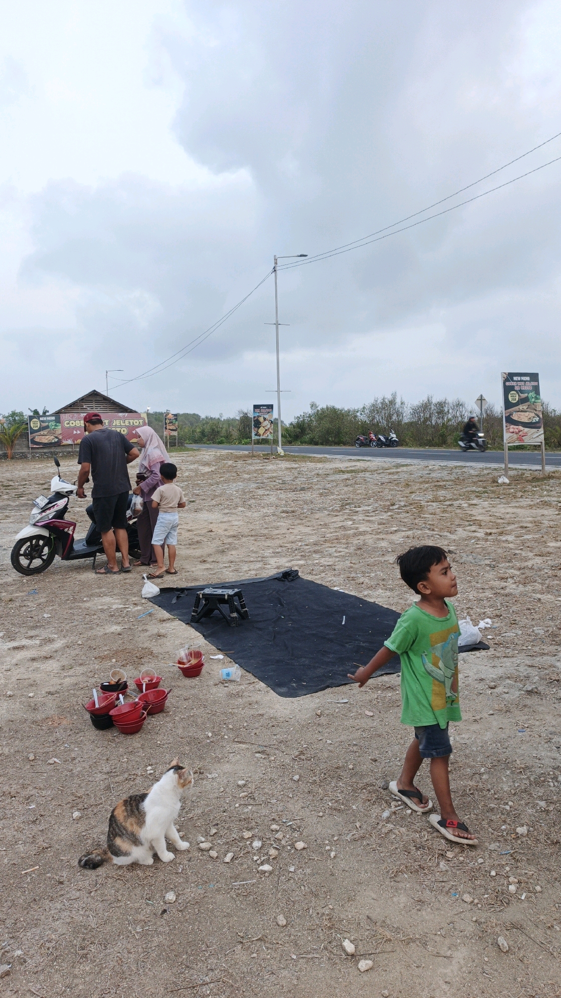 In Pangandaran, Indonesia, on an overcast day, a family is gathered near a roadside establishment. A man in a dark t-shirt and shorts stands beside a white and pink scooter, interacting with a woman wearing a pink hijab and a light blue outfit. A young boy in a white t-shirt and blue shorts stands to their side, facing away from them. In the foreground, a young boy in a green t-shirt and denim shorts looks to the right, with his arm extended. A calico cat sits on the dusty ground to the left, looking to the right. Scattered around are several red buckets, some containing what appears to be food, and a black tarp lies on the ground with a small black stool on it. In the background, a paved road is visible with several motorcycles driving by. Billboards advertising food are placed along the roadside, and a simple wooden building with a thatched roof is situated behind the family. The environment is characterized by dry, sandy ground and sparse vegetation.