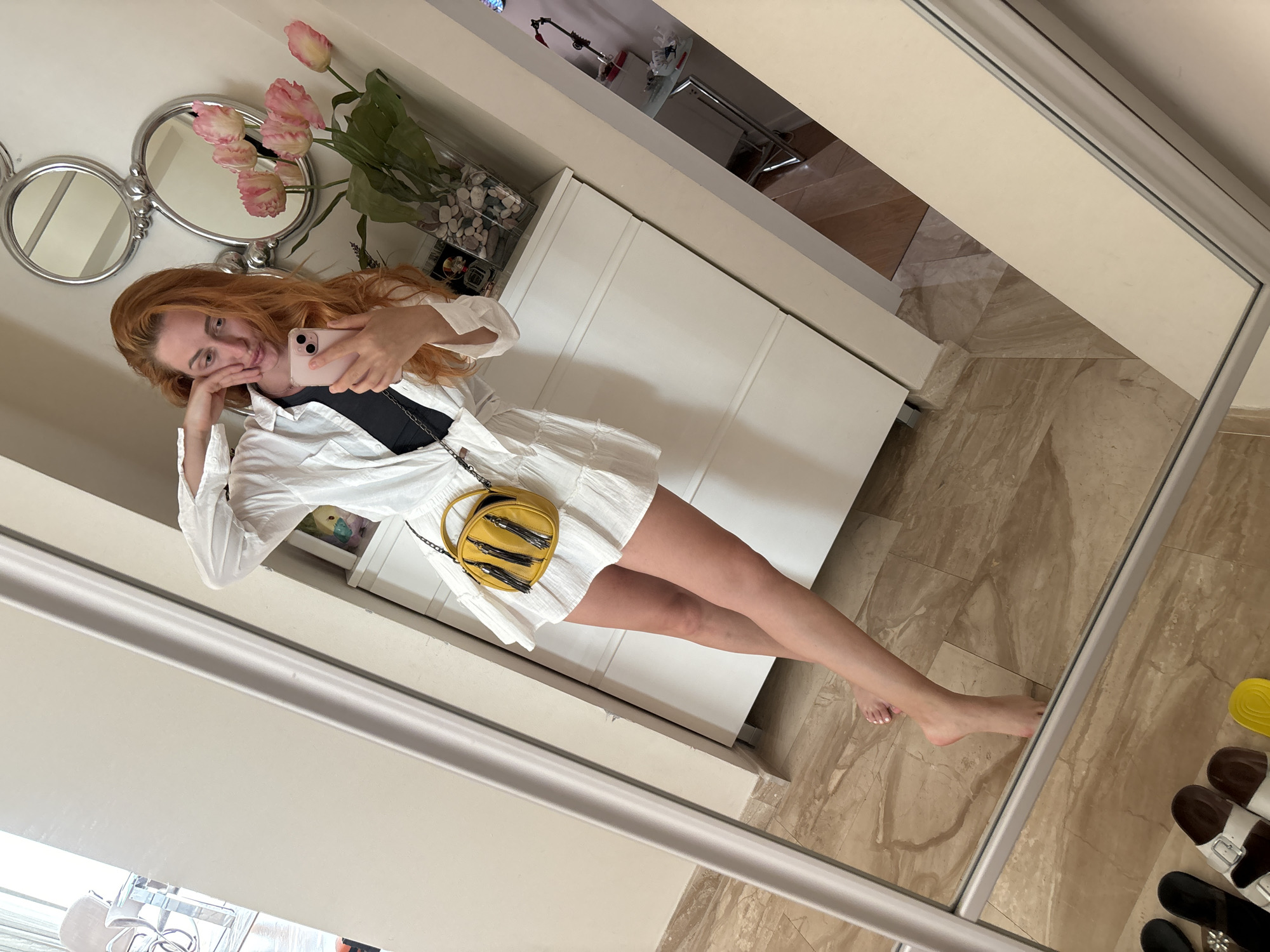 A woman with red hair is taking a mirror selfie in what appears to be a bedroom or dressing area in Budva, Montenegro. She is wearing a white button-down shirt over a black top and a white ruffled skirt. She has a yellow crossbody bag with black tassels. She is leaning her hand on her face and holding a pink smartphone. In the background, there are decorative mirrors, a vase of pink flowers, and a white dresser. The floor is tiled with a marble pattern. Several pairs of sandals are visible in the foreground reflection.
