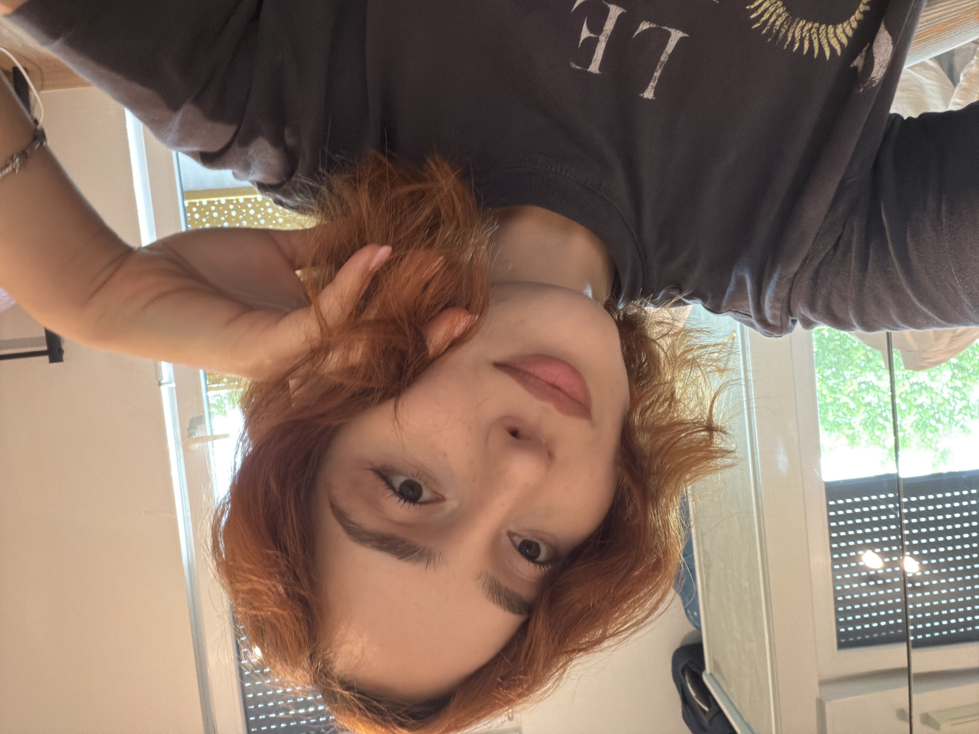 A young woman with shoulder-length auburn hair is taking a selfie in what appears to be an indoor setting, possibly a room in Strasbourg, France, given the context. She is wearing a dark grey t-shirt with some graphic elements visible on the front. Her left hand is resting on her cheek, and she is looking towards the camera with a soft smile. The background shows a window with blinds and trees visible outside, suggesting a daytime scene. A doorway with a handle and a white frame is also visible. The lighting is soft, indicating natural light from the window.