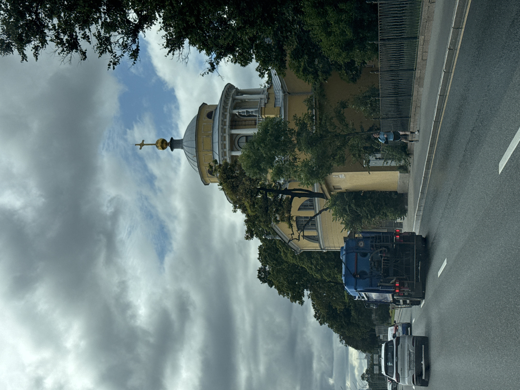 The image depicts a view from a vehicle on a road in Saint Petersburg, Russia, on a cloudy day. In the background, a yellow building with a domed roof topped with a cross is visible, surrounded by lush green trees. A man is jogging on the sidewalk next to the building. In the foreground, the road features multiple lanes and vehicles, including a white SUV and a large blue truck. The sky is heavily overcast with dramatic clouds, typical of Saint Petersburg's climate. The overall scene captures a moment of urban life with architectural and natural elements.