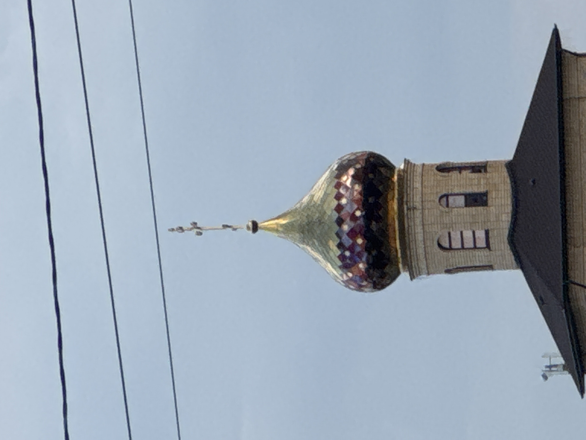 A tall, golden, onion-shaped dome topped with a cross stands against a light blue sky. The dome is adorned with a mosaic of colored tiles in shades of purple, gold, and red. Below the dome is a cylindrical tower with arched windows, constructed from pale yellow brick. The tower is capped with a dark, sloping roof. Three power lines stretch horizontally across the upper portion of the image. The lower part of the image shows the peak of a dark roof.