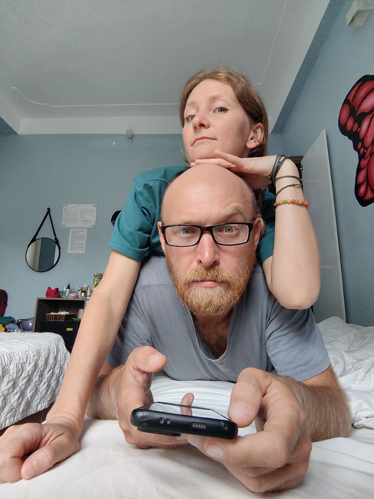 Two individuals, a male and a female, are positioned on a bed. The male lies prone, holding a mobile phone in his hands. He has a reddish beard and is wearing glasses and a grey T-shirt. The female is positioned behind him, with her left hand resting on his head and her right arm extended to the side. She is wearing a green T-shirt and several bracelets on her right wrist. The room has pale blue walls. A butterfly decal is visible on the right wall, and a round mirror and documents are on the left wall above a small storage unit next to another bed.
Given the presence of Vietnamese currency, items, and the building and room styles, the location is likely in Vietnam. The city of Bai Chay Ward is inferred due to the general style of the room matching what is commonly found in this type of accommodation.