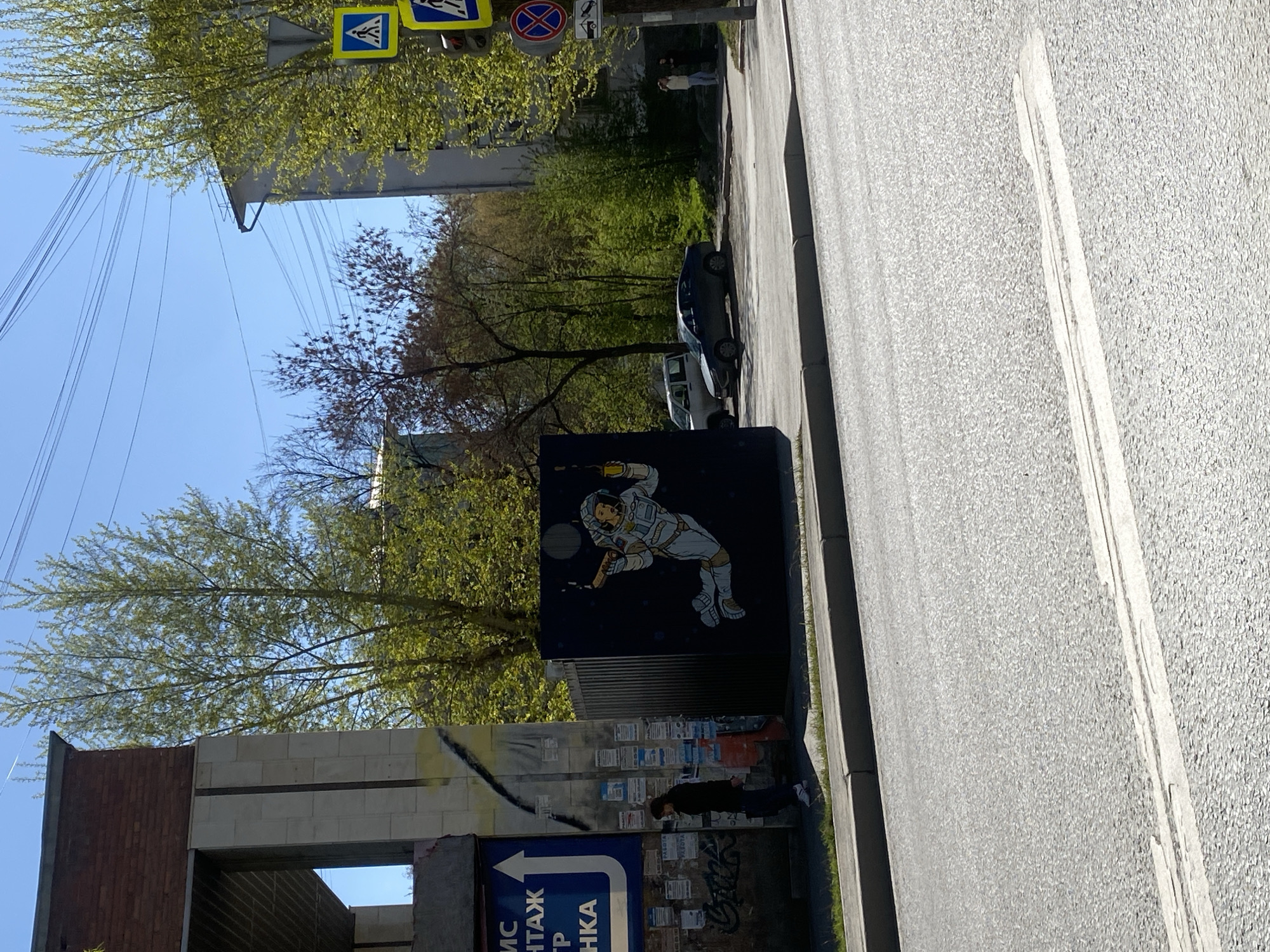 The image shows a street scene in Yekaterinburg, Russia. A partially constructed or dilapidated building stands on the left side of the frame. On a wall attached to this building is a large mural depicting an astronaut playing tennis. A man stands in front of the wall, seemingly examining flyers posted there. A street runs along the right side of the frame with parked cars visible in the background. Overhead, trees with green foliage are visible against a blue sky. In the upper portion of the image, traffic signs are mounted on poles.