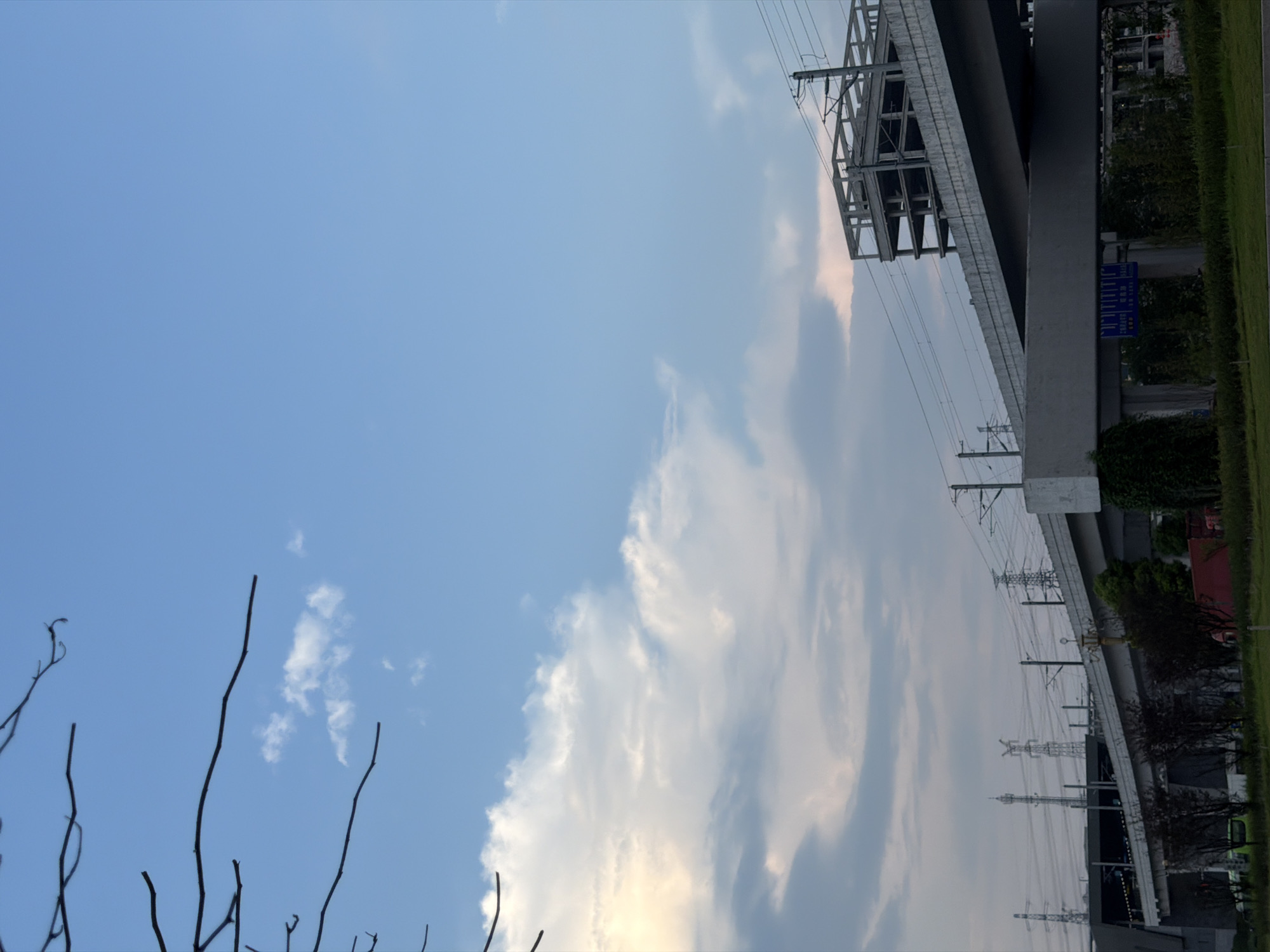 The image depicts an elevated roadway and railway structure under a partly cloudy sky. In the foreground, there is a bare tree branch extending from the upper left corner. The roadway, constructed of concrete, curves into the frame from the right, with power lines running along its edge. Below the roadway, a railway track is visible with overhead power lines. In the distance, several power transmission towers are scattered across the landscape. The sky transitions from a clear blue at the top to a gradient of lighter hues with scattered clouds towards the horizon, where some sunlight is breaking through. The overall scene suggests an urban or semi-urban environment, possibly in Jiangjin, China, given the presence of elevated infrastructure and power lines typical of such locations. There are no visible people in the image.