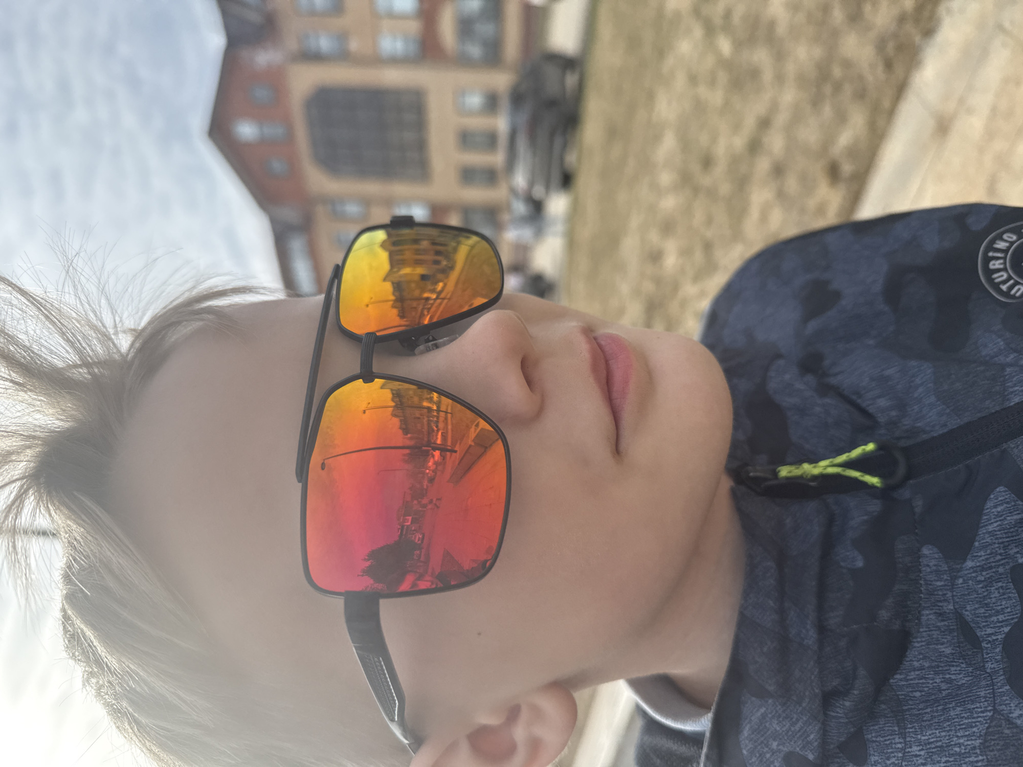 A young male, estimated to be between 8 and 12 years old, is depicted in a close-up, slightly low-angle outdoor photograph. He has light-colored hair and wears black-framed sunglasses with highly reflective, reddish-orange lenses. He is dressed in a dark, camouflage-patterned jacket featuring a neon yellow zipper pull. The background is out of focus. Directly behind him, a multi-story building with a red roof and light-brown facade, containing numerous windows and a prominent ground-level archway, is visible. This building is identified as the Youth Center 'Horizon' at ul. Pushkinskaya, 24, Dmitrov, Russia. The reflections in the sunglasses display a clear image of a street scene, including vehicles, streetlights, and other buildings, indicating an urban setting. The ground in the foreground is dry, consisting of light brown earth or sparse grass, with a paved edge partially visible. The sky is bright with visible cloud patterns. The subject's head is turned slightly to the right, and his gaze follows this direction.