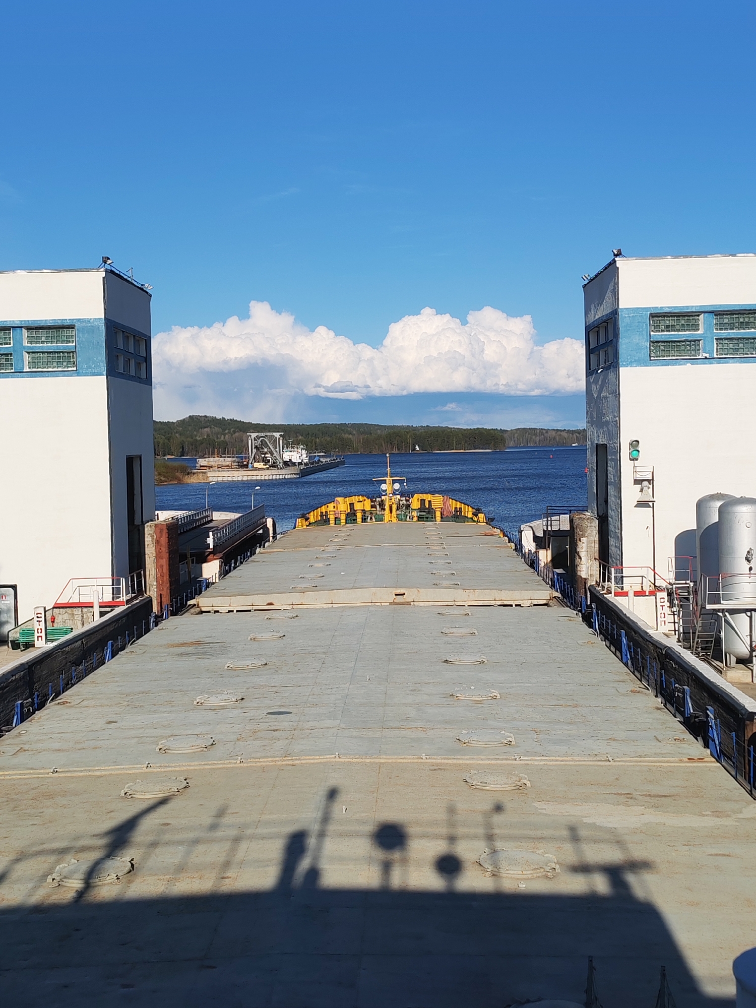 This is an outdoor shot of a canal lock in Белоусово, Russia, taken on a bright day with scattered clouds. The view is from within the lock, looking out toward a body of water with forested shoreline. A wide, flat-topped vessel occupies the lock. The vessel has a gray, metallic surface marked with round hatches and shadowed figures, with yellow equipment towards the far end. The lock is flanked by tall, white concrete structures with blue accents, including windows and horizontal stripes. A bridge and another vessel are visible on the other side of the waterway in the distance.
