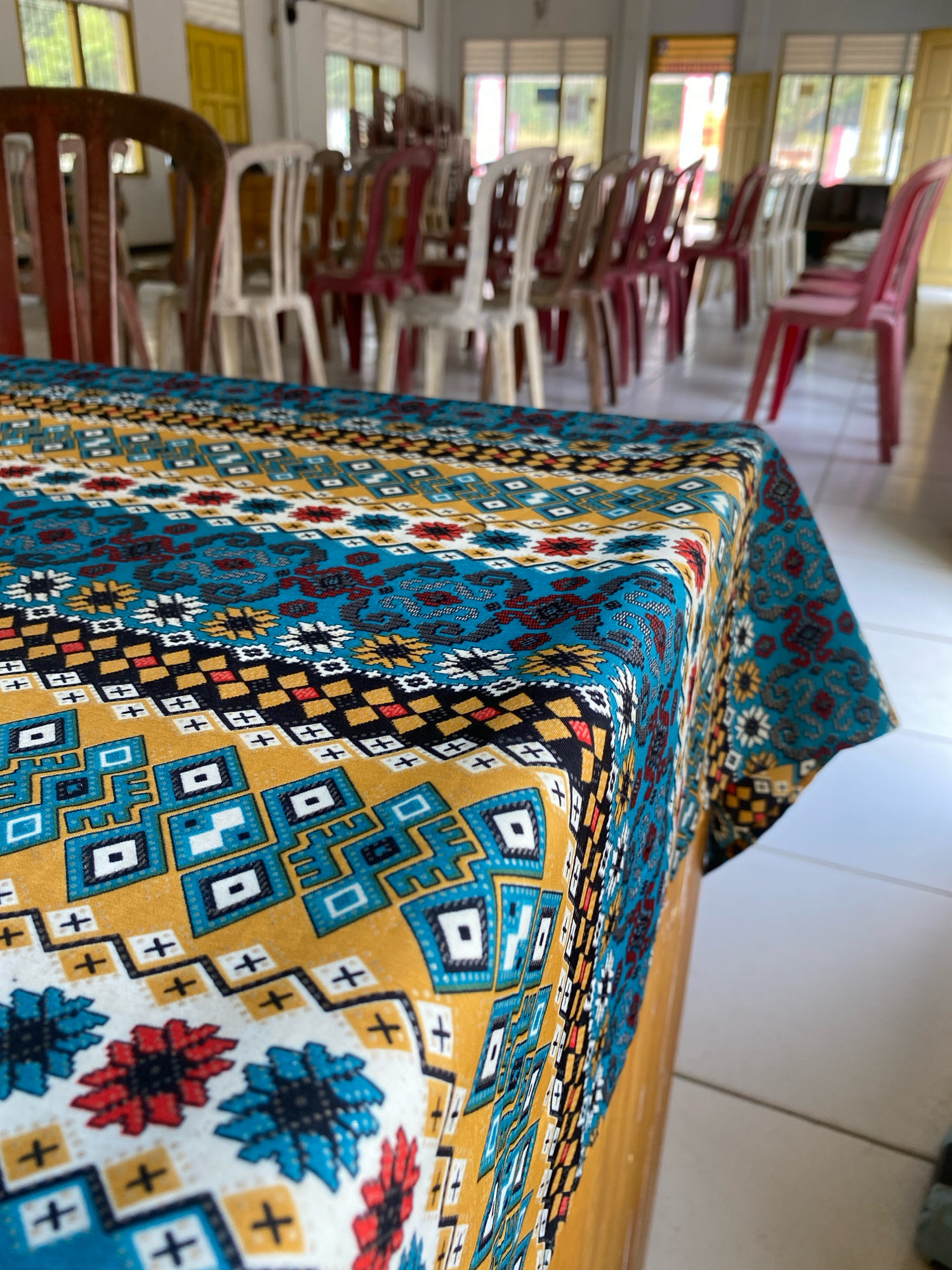 The image shows a close-up of a patterned tablecloth draped over a wooden table. The tablecloth features a vibrant, geometric design with colors of teal, yellow, white, red, and black. The background is blurred but appears to be an indoor setting with rows of plastic chairs in various colors, including white, red, and pink. The chairs are arranged as if for a gathering or event. The environment suggests a community hall or a similar public space. The location is specified as Mojo Pahit, Indonesia.