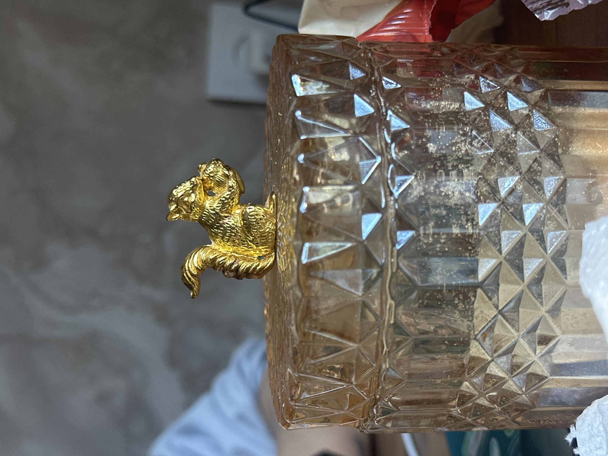 The image displays a close-up, rotated view of a decorative golden figurine attached to a clear, textured glass container. The golden object is an intricately sculpted animal figure, possibly a lion or a mythical creature, featuring detailed fur or a mane and a curled tail, with a highly reflective surface.
The glass container, to which the figurine is affixed, is made of clear or lightly amber-tinted glass and features a prominent, repeating diamond-cut or faceted pattern across its surface. Light reflects off these facets, creating bright highlights. Inside or on the surface of the glass, some residue or fine dust particles are visible. A small piece of red material, possibly plastic or fabric, is partially visible near the top edge of the container, and some white fabric or tissue is seen along the bottom right edge.
The background is blurred, showing a light grey wall with an electrical outlet visible in the upper left. A person's arm or shoulder, wearing a light-colored shirt, is faintly discernible in the lower left portion of the background. The scene appears to be an indoor setting under ambient light. No specific activity is taking place, and there is no visible text.