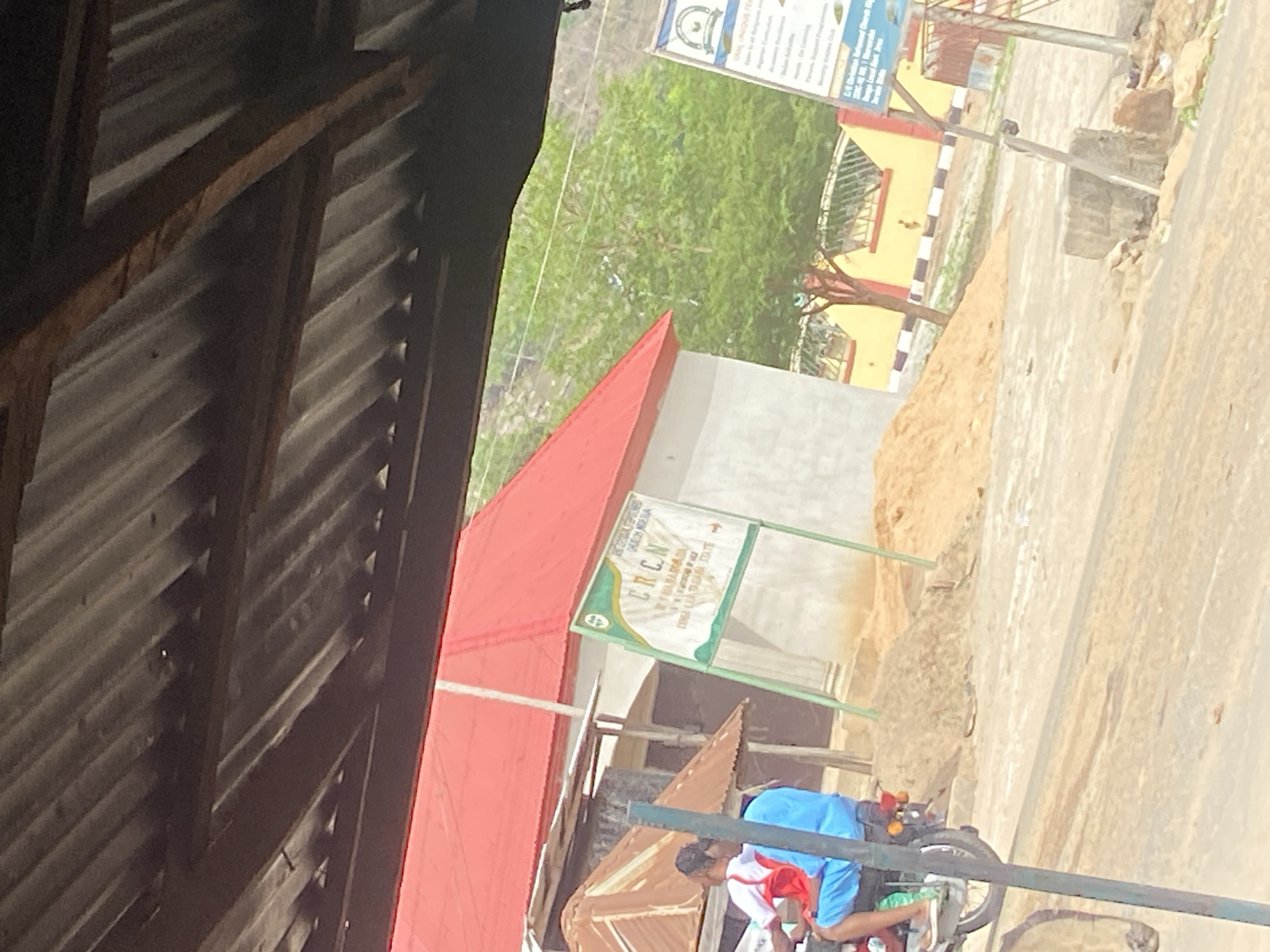 The image presents a bright, sunny daytime scene, likely in Mararaba, Nigeria, viewed from beneath a corrugated metal roof supported by wooden beams. The roof structure occupies the upper and left portions of the foreground, creating deep shadows.
In the mid-ground, a building with a prominent red corrugated roof stands on the left. Adjacent to it, a green-framed sign is clearly visible. The text on this sign identifies it as belonging to the "NATIONAL EPISCOPAL COMMISSION FOR DOCTRINES, ECUMENISM AND INTER-RELIGIOUS DIALOGUE NIGERIA" and "E.R.C.N" (Reformed Church of Nigeria), specifying the location as "KASUWAN MEKA, KM 10, LAFIA ROAD, MARARABA, KARU LGA, NASARAWA STATE".
To the right of the red-roofed building, a pile of sand sits beside a dusty, light-colored road or path. In the lower right corner, two individuals are partially visible on a motorcycle; one wears a blue top, and the other a red and white top.
The background features lush green foliage, suggesting trees or bushes. Another building, with a yellow facade and a black and white striped curb, is visible further back. In the upper right, a second large sign is partially seen, listing "UNIQUE FEATURES" such as "Open to all backgrounds," "Nigerian Curriculum," "Academic Excellence," "Modern Facilities," "Skills Acquisition," "e-Learning," "Parents Support Programme," "Excursion & Field Trips," "Debate & Quiz," and "Mentoring & Guidance Club." The scene depicts an everyday street environment in a local community.