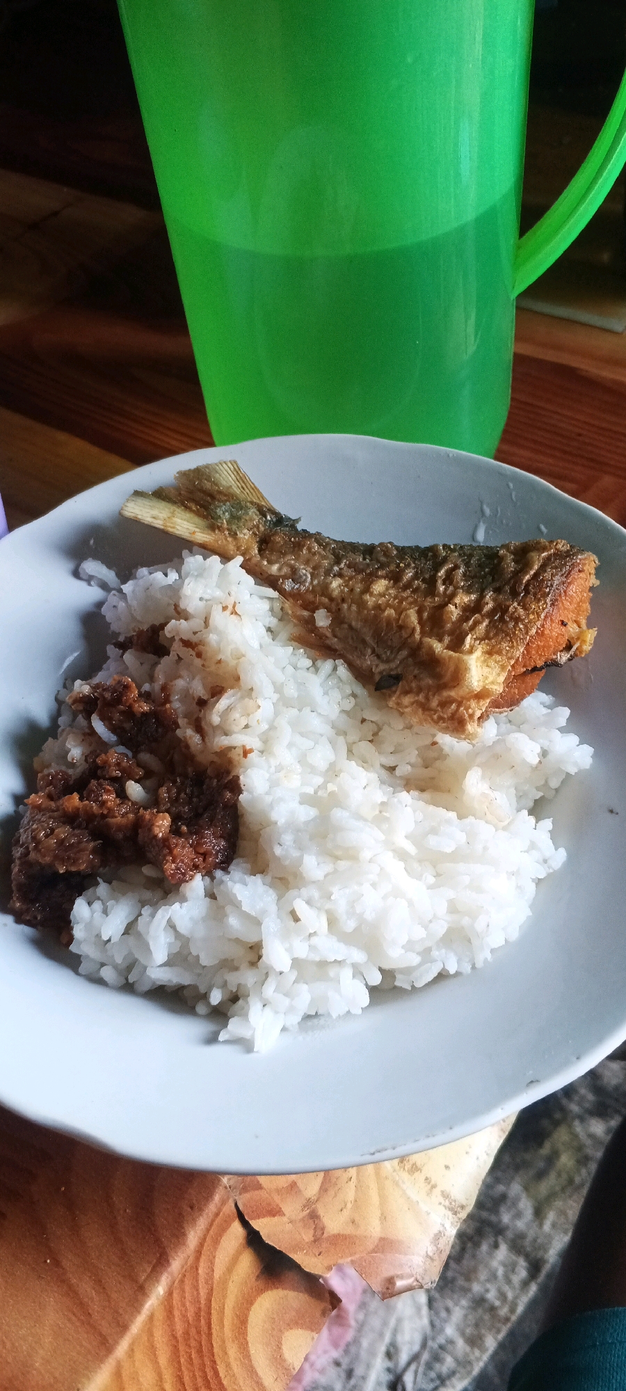 A plate of rice with fried fish and a dark-colored topping sits on a wooden table. A green plastic jug filled with liquid is visible in the background, also on the table. The table appears to be made of wood planks. The lighting suggests an indoor setting, possibly a dining area. The image is focused on the food on the plate. There is no indication of the specific location within Penanggungan, Indonesia.