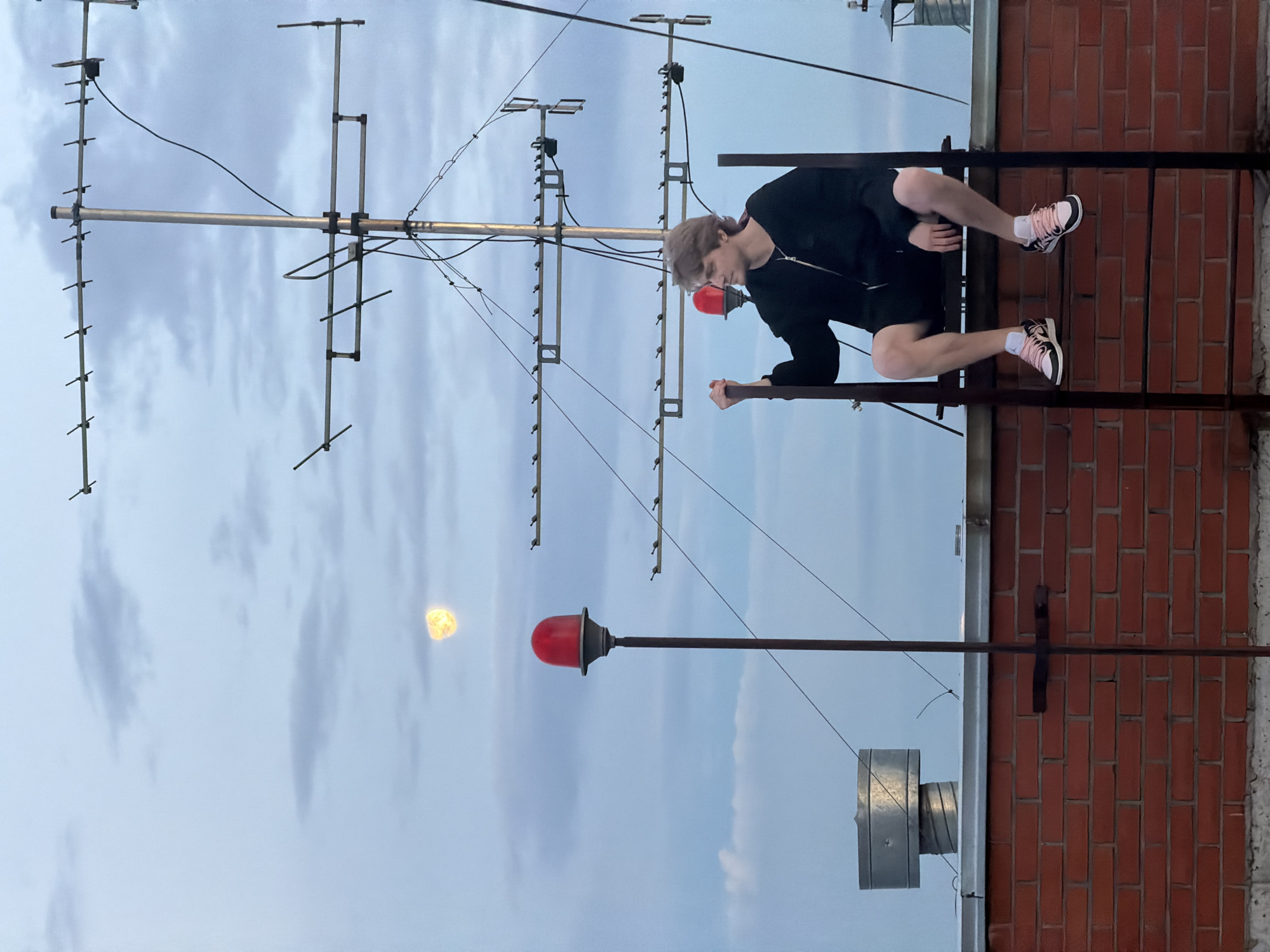 A young man with light brown hair is sitting on a metal ladder structure on a brick rooftop in Khimki, Russia. He is wearing a black zip-up hoodie, black shorts, and pink and white sneakers. He is looking to his right, away from the camera. In the background, there is a clear sky with some clouds and a bright, full moon. There are also several antennas and a red warning light visible on the rooftop. The overall scene suggests an urban setting, possibly on a building overlooking the city.