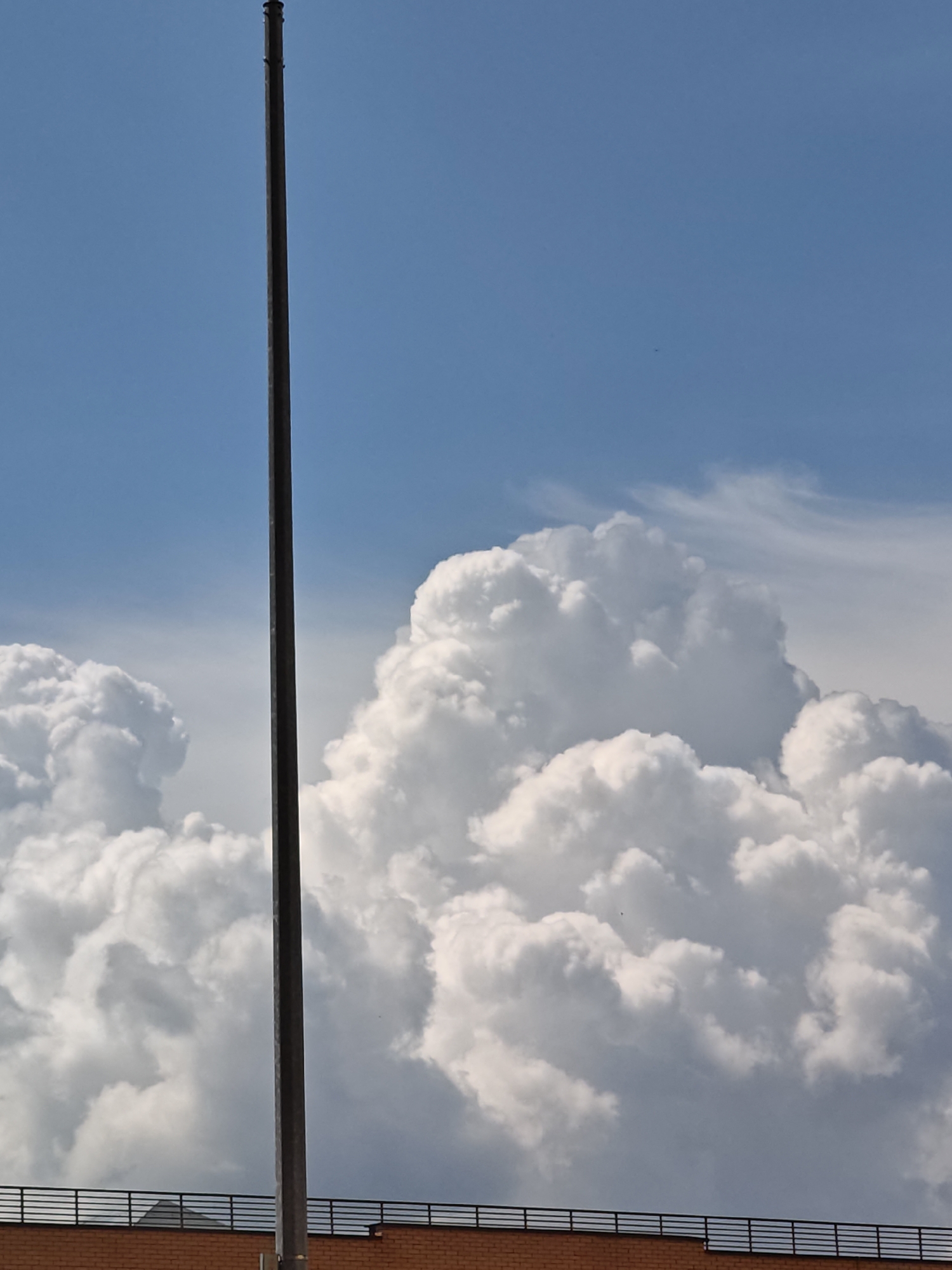 A tall, dark, vertical pole stands against a bright blue sky filled with fluffy white cumulus clouds. The bottom of the image shows the top of a brick building with a dark railing. The scene appears to be an outdoor setting, possibly in the city of Shcherbinka, Russia, given the context. There are no people or animals visible in the frame. The light suggests it is daytime.