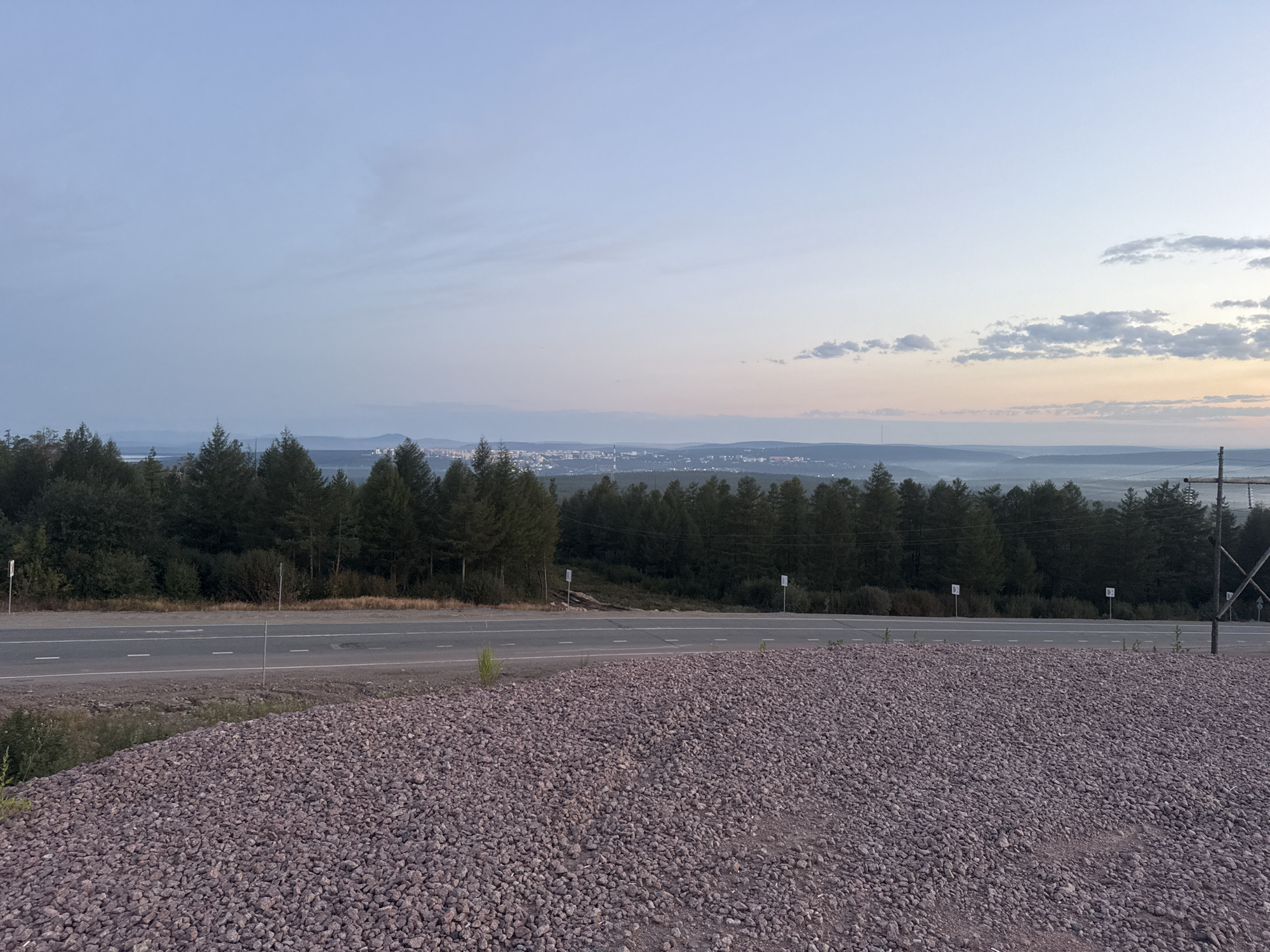 The image depicts a roadside scene at dusk, overlooking a distant city. In the foreground, a large pile of reddish-purple gravel occupies the lower half of the frame. A paved road with white lane markings runs horizontally across the middle of the image, separating the gravel from a dense line of dark green coniferous trees. Beyond the trees, the landscape recedes into rolling hills and a hazy valley. In the far distance, a city skyline is visible, with lights twinkling amidst the fading daylight. The sky above is a gradient of soft blues and pinks, with scattered clouds. The location is identified as Serebryany Bor, Russia. The scene is devoid of any people or visible activity, presenting a quiet, natural landscape with a man-made road and a distant urban settlement.