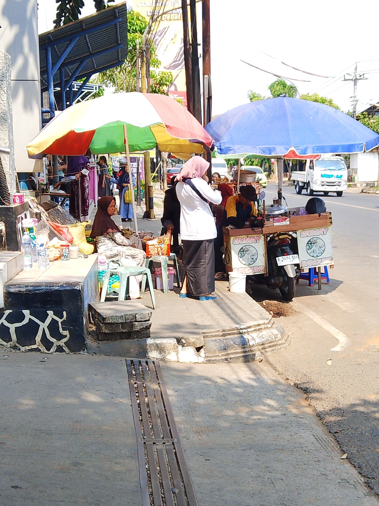 The image depicts a street scene in Potroyudan, Indonesia, with several people engaged in selling and purchasing goods. A woman wearing a brown hijab and a patterned dress sits on a raised concrete ledge, appearing to be tending to her stall. In front of her are various items, including bottles of water, packaged snacks, and containers. Adjacent to her stall, a food cart with the words "SIOMAY BANDUNG" displayed prominently on its sides is set up. A man in a yellow and blue shirt is visible behind the cart, seemingly preparing food. Another woman, wearing a pink hijab and a white top, stands facing the cart, possibly as a customer. The food cart is mounted on a motorcycle, and a blue stool is placed beside it. The scene is shaded by two large umbrellas, one red, yellow, and green, and another blue. In the background, a white truck is parked on the road, and a yellow car is also visible. The environment appears to be an outdoor market or roadside vending area, with a paved street and sidewalk. The lighting suggests it is daytime.