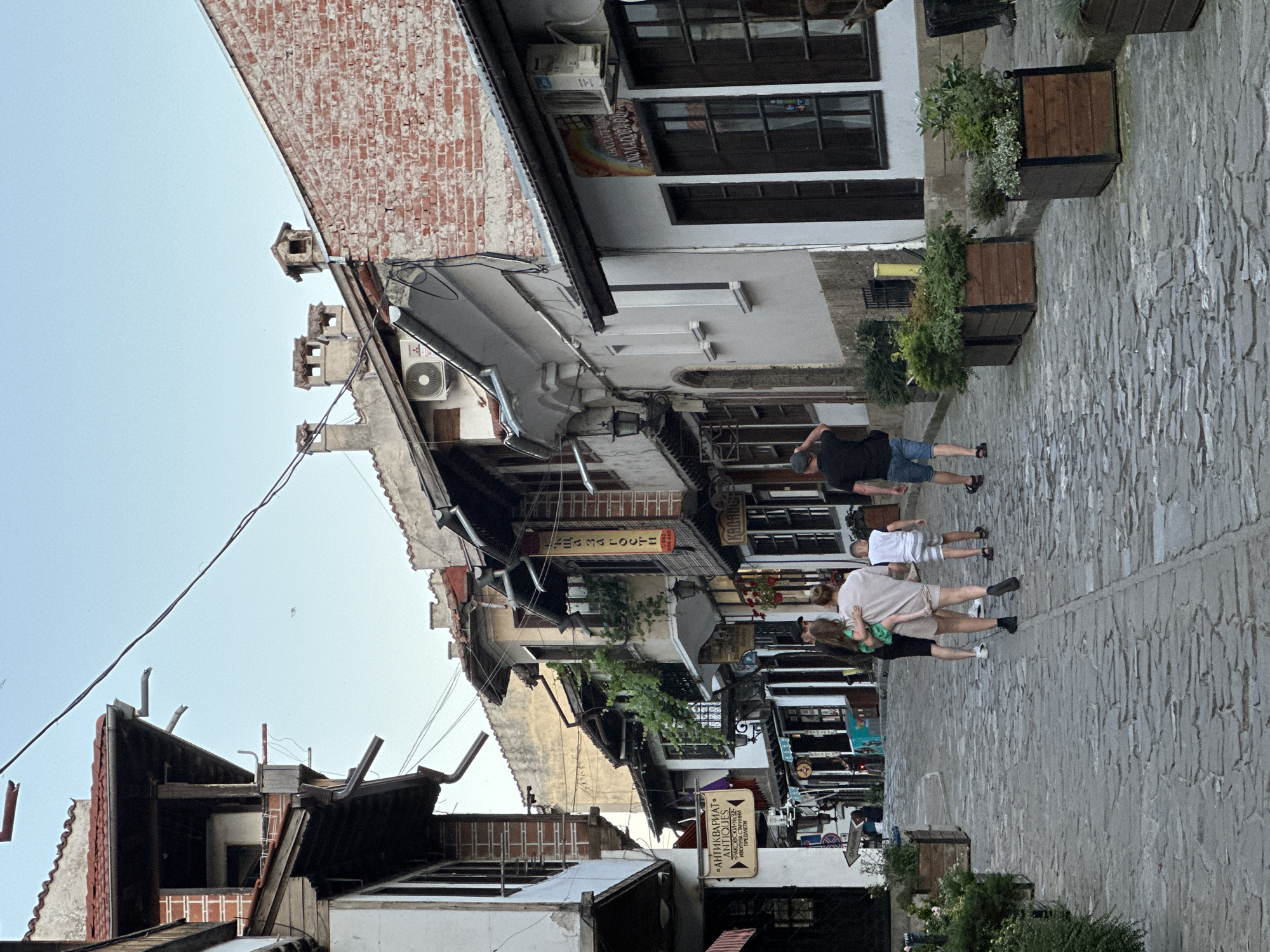 The image shows a cobblestone street in Veliko Tarnovo, Bulgaria. The street is lined with old, multi-story buildings made of stone and brick, some with terracotta tiled roofs. Many of the buildings have wooden balconies and are adorned with plants. Several signs are visible on the buildings, including one for "ANTIQUARIAT ANTIQUES" and another with Cyrillic text that appears to advertise a chocolate shop. The sky is a light blue, suggesting it is either early morning or late afternoon.
There are several people walking on the street. In the foreground, a family is walking away from the camera. A woman is carrying a young child, and a boy is walking beside them. Further down the street, other individuals can be seen walking. Planters with green foliage are placed along the sides of the street. The overall scene depicts a quaint, historic street in a European city.