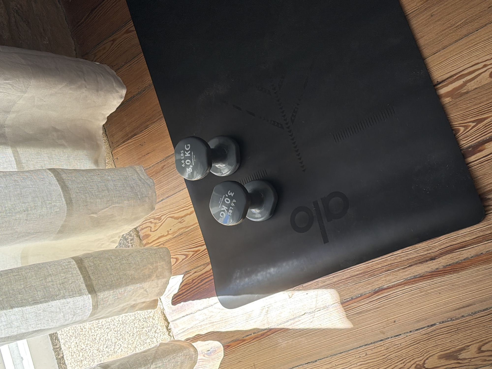 A high-angle, close-up shot shows a black yoga mat on a wooden floor. Two gray dumbbells, each labeled "6.6 LBS 3.0 KG", are placed on the mat. Natural light streams in from the left, creating shadows and highlighting the texture of the wooden floor and the mat. In the background, a sheer white curtain hangs, also illuminated by the natural light. The scene appears to be indoors, likely in a domestic setting. There are no people or animals present in the image. This image does not contain any information that would place it specifically in Santiago De Compostela, Spain.