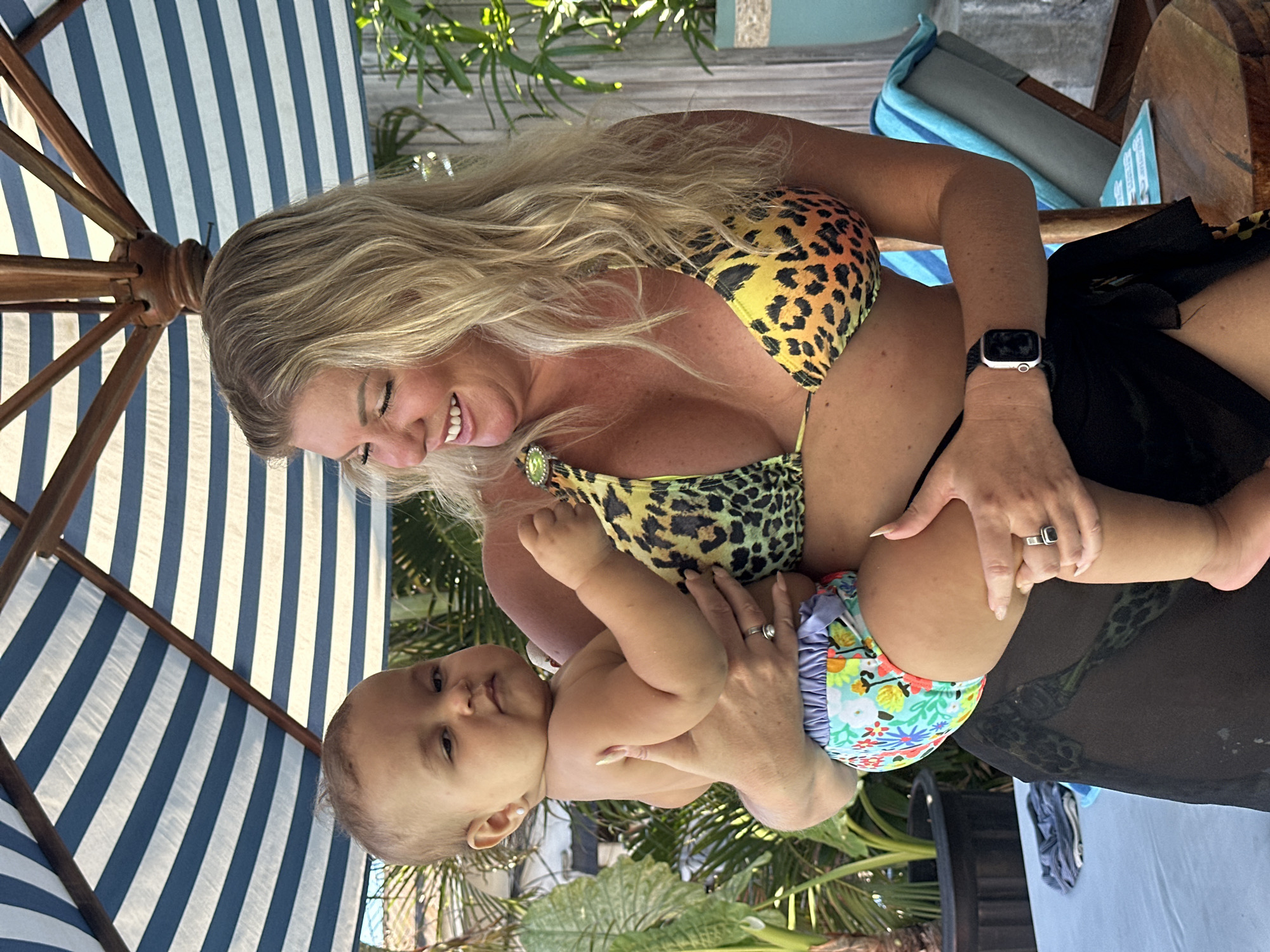 A woman in a leopard print bikini is holding a baby in a floral diaper. They are under a blue and white striped beach umbrella. The location is likely in Kuta, Indonesia, as this is a popular beach destination. There are green plants and trees in the background. The woman is wearing a watch on her left wrist and rings on both hands.