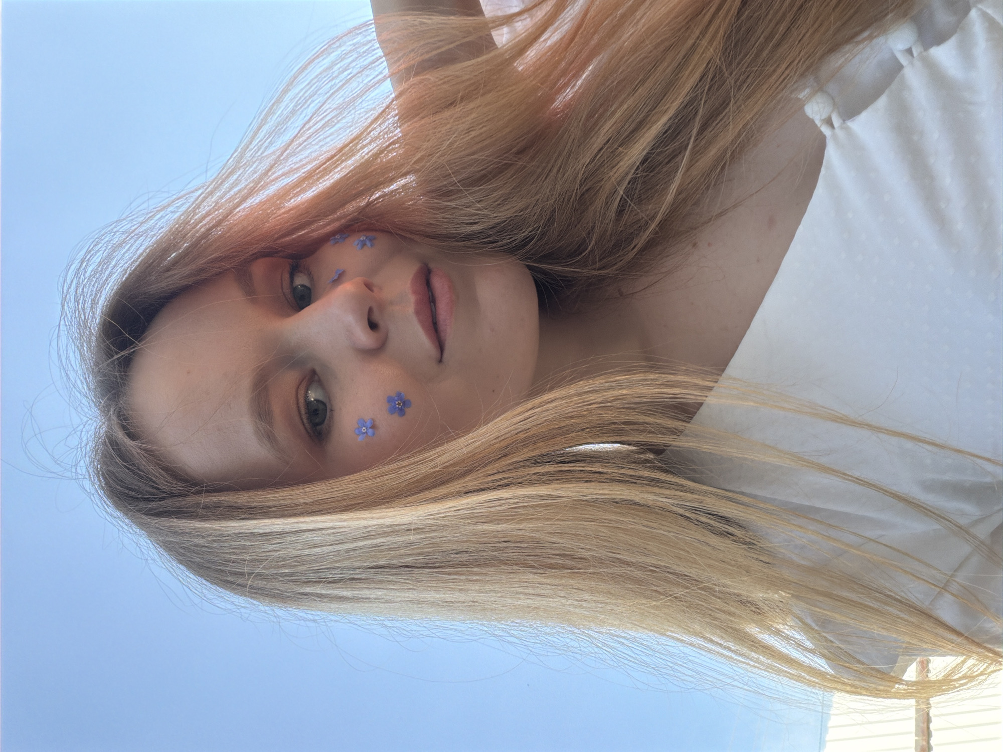A woman with long blonde hair is facing the camera, her face adorned with small blue flowers near her eye. She is wearing a white blouse. The background shows a clear, pale blue sky. The location is Кадряково, Russia.