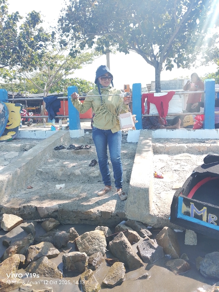 This image depicts an outdoor scene in Limbangan, Indonesia, under bright sunlight, possibly near a waterfront or riverbank. A female individual stands centrally in the foreground, directly facing the camera. She wears a blue bucket hat, sunglasses, a long-sleeved light olive-green top, blue jeans, and light-colored flat shoes. She carries a light-colored shoulder bag and holds a thin, metallic object horizontally with both hands. In her right hand, she also holds a clear cup containing an amber liquid.
Behind the primary subject are concrete steps leading downwards, on which several pairs of dark sandals are placed. Further back, blue-painted concrete posts form a barrier. To the right of the main subject, a female figure in a red shirt is visible from the chest up, seated or leaning against the blue barrier, with a red garment draped over it. To the left, a partially obscured figure in a red shirt is also visible behind the barrier. Large trees with green foliage stand in the mid-ground and background, under a bright sky exhibiting significant sun flare. The immediate foreground and lowest visible area consist of numerous large, irregular rocks partially submerged in wet sand or mud. On the far right, a dark object resembling a portion of a boat or decorative structure, featuring "MR" painted in blue and yellow, is partially visible at the water level.