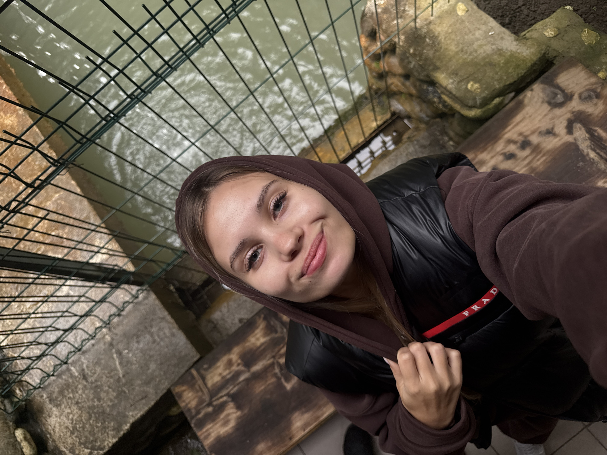 A selfie angled downwards shows a young woman in Tsabal, Abkhazia. She is wearing a brown hoodie and a black Prada vest. She smiles slightly at the camera. In the background are a metal grate, water, stone, and weathered wooden planks.