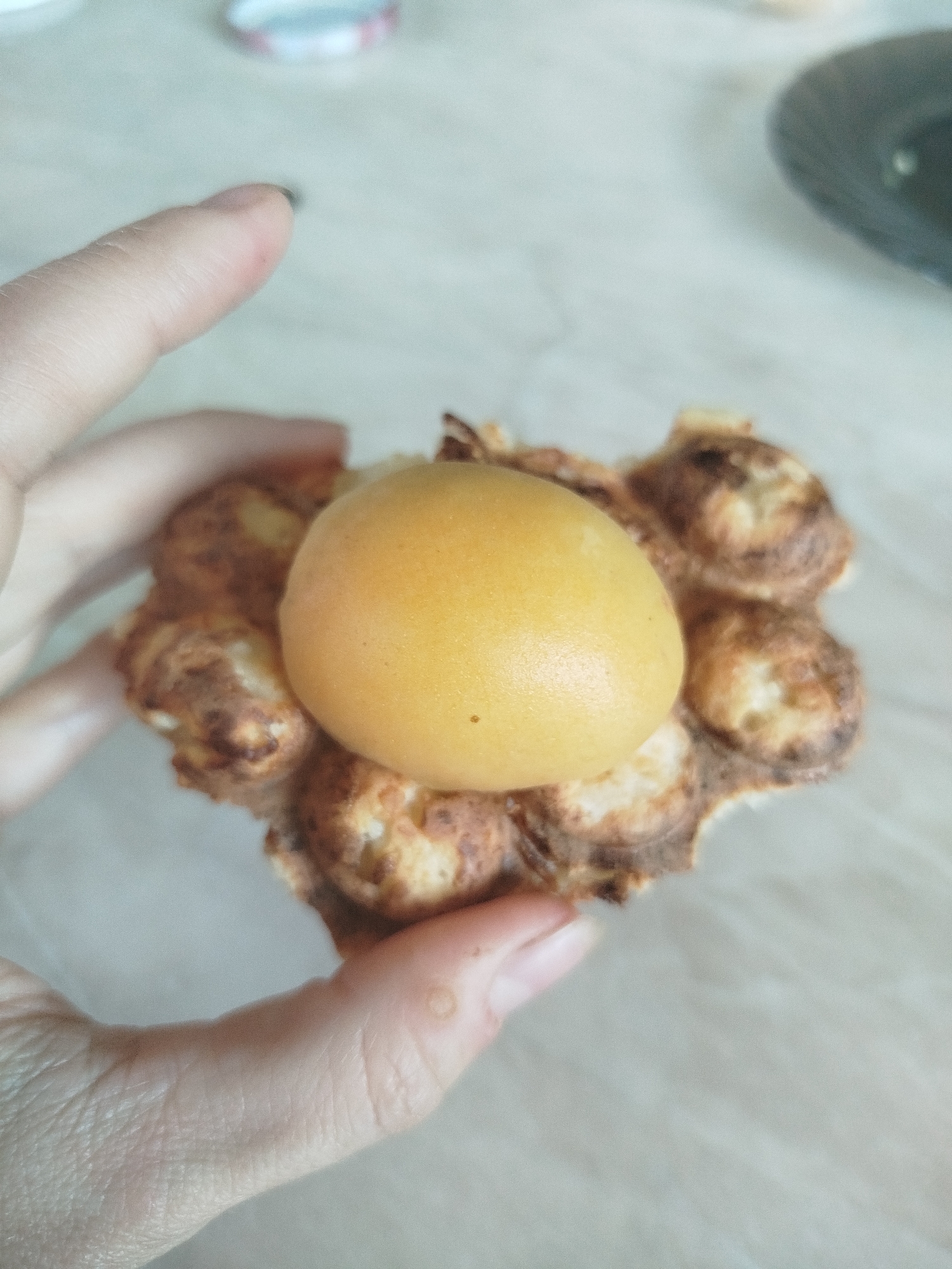 A close-up, top-down shot shows a person's hand holding a bubble waffle topped with a single, bright yellow apricot. The waffle is golden brown and slightly charred in places, with a textured, bubbly surface. The apricot is smooth and round, positioned centrally on the waffle. The background consists of a light-colored, possibly marble-patterned countertop. The image does not provide any information about the location in the city of Zavodoukovsk, Russia.