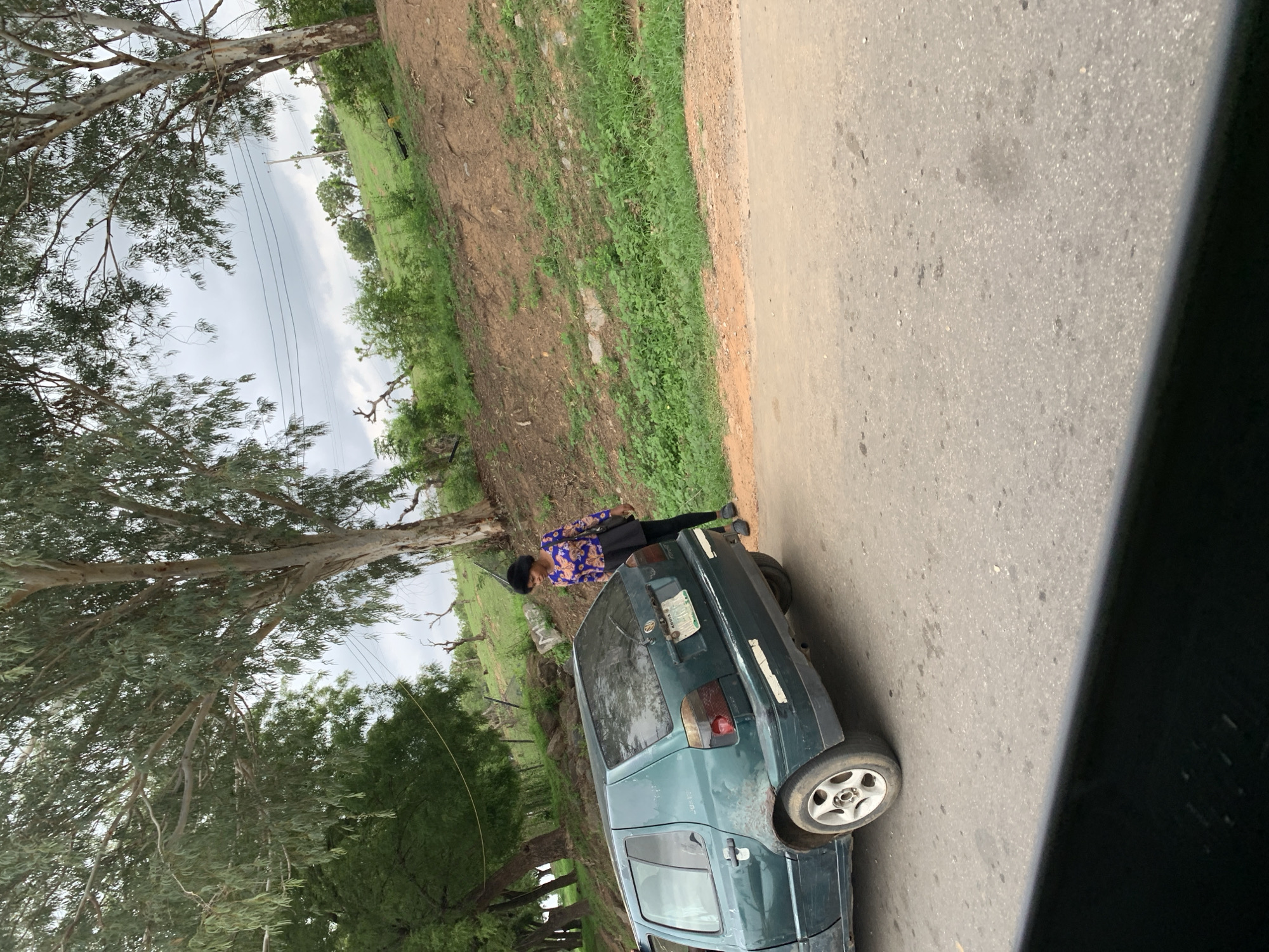 The image depicts a roadside scene, likely in a rural or semi-rural area, under an overcast sky. The photograph is oriented vertically, rotated 90 degrees counter-clockwise.
In the mid-ground, a dark green or blue sedan is parked on the side of an asphalt road, facing away from the viewer. The car shows signs of wear, including white tape on its bodywork near the rear lights. The right rear wheel has a silver five-spoke rim. A license plate is affixed to the rear of the car, but its text is illegible.
Standing near the driver's side rear of the car is a person, dressed in a long-sleeved top with a blue, orange, and black pattern, black trousers or a long skirt, and dark shoes. They also wear a dark head covering or hat. The person is looking towards the left side of the frame, away from the camera.
The road, visible on the right, is made of asphalt with a textured surface. To the left of the road and the car, the terrain consists of a mix of green grass and patches of bare brown earth. Further into the background, beyond a line of fencing or poles, lie expansive green fields dotted with more trees, characteristic of a verdant landscape. Large trees with light-colored bark and abundant foliage dominate the left side of the frame, their branches reaching across the scene. Thin power lines are visible against the cloudy sky above the trees.
The scene suggests daytime under cloudy weather. The setting, with its specific vegetation and road conditions, aligns with the provided location context of Gwaska, Nigeria. No specific activity beyond standing near a parked car is discernible. No legible text is visible other than the unreadable car license plate.