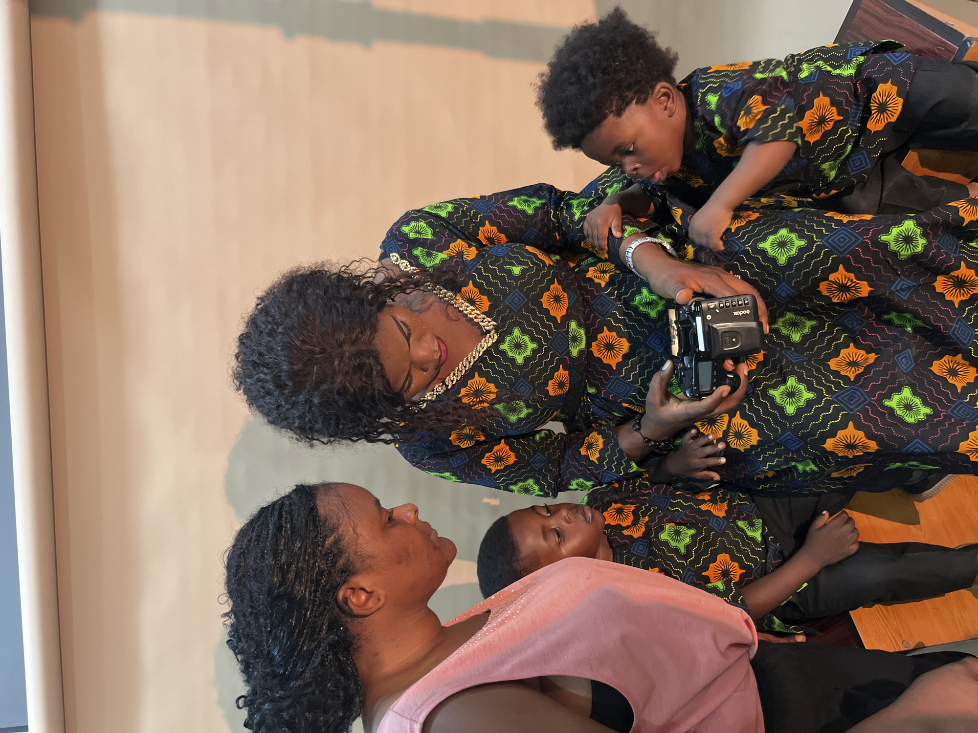 In this vertical, eye-level shot, a group of three individuals are positioned in a studio setting with a seamless beige backdrop. The subjects are an adult female and two young boys, all of whom appear to be of African descent. They are dressed in matching patterned garments featuring a striking design of orange, green, and blue floral motifs on a black background, accented with geometric patterns in pink and blue.
The adult female, with dark, curly hair and prominent gold jewelry, is looking down with her eyes closed. She is holding a black Kodak camera in her right hand, with a young boy's hand resting on hers. This boy, also in the matching attire, is leaning over her, looking intently at the camera.
In the foreground, another young boy, wearing a pink top over black clothing, looks up towards the adult female. His expression is calm and engaged. The overall scene suggests a candid moment, possibly during a photo session or a shared activity involving the camera. The lighting is soft and diffused, indicative of indoor studio lighting, and there are no clear indicators of the time of day or weather conditions. The setting appears to be a studio, given the backdrop and lighting, and the presence of a wooden surface in the lower right corner.