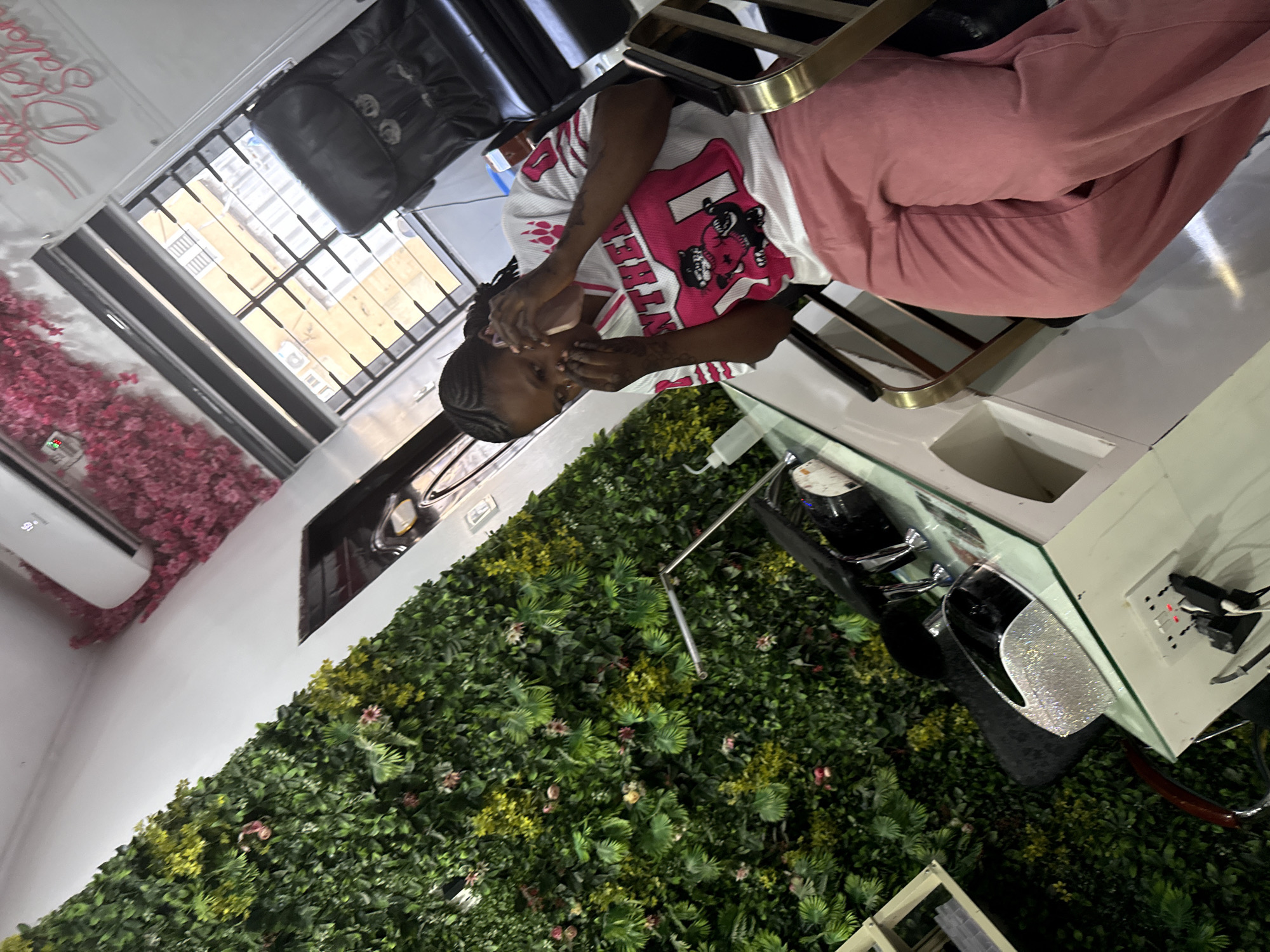The image shows a person seated in a chair inside what appears to be a beauty salon or similar establishment. The person is wearing a white t-shirt with a pink graphic design and pink pants. Their hands are raised to their face. Behind the person, there is a wall covered in green foliage with some yellow flowering plants interspersed. A window is visible in the upper left corner, showing buildings outside. Further to the left, a wall is adorned with pink floral decorations. The salon setting is evident from the salon chair and surrounding equipment. The location in Lugbe, Nigeria cannot be verified with this image.