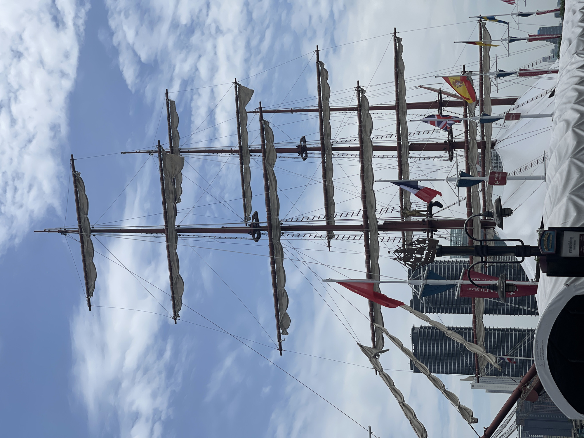 This is a medium-angle shot of a large, multi-masted sailing ship docked in the Chao Phraya River in Bangkok, Thailand. The ship's sails are furled, and its rigging is intricate and extensive. Several national flags, including those of France, the United Kingdom, Spain, and possibly Germany and Sweden, are visible on display. In the background, modern high-rise buildings characteristic of Bangkok's cityscape rise into a sky filled with scattered clouds. The ship appears to be a tourist attraction or event space, with a white tent-like structure partially obscuring the lower deck. The image captures a juxtaposition of historical maritime architecture against a backdrop of contemporary urban development.