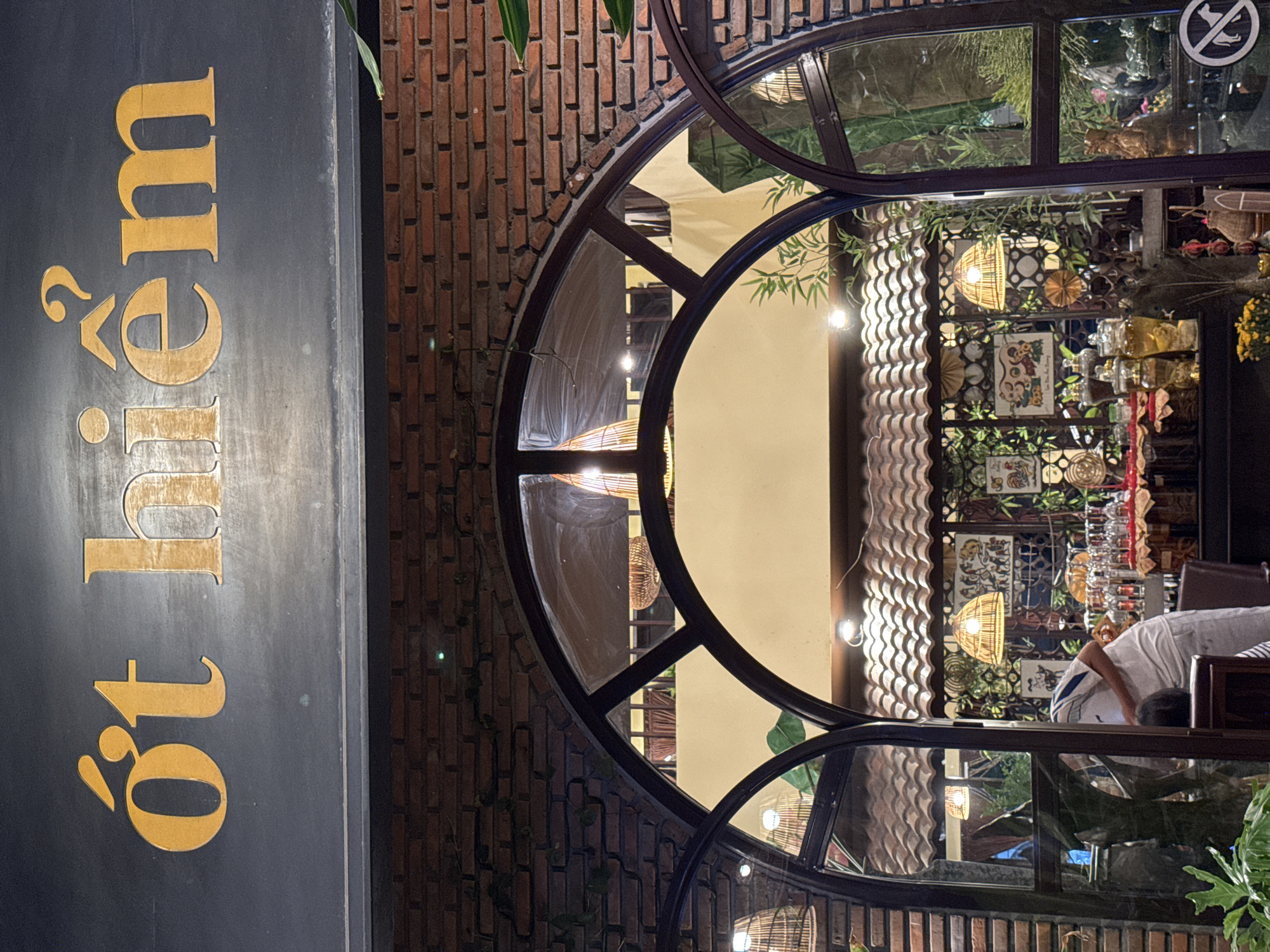 The image shows the exterior of a building at night. A dark-colored sign with gold lettering is visible on the left, with the words "ốt 19" appearing vertically. The building features a large, arched window with dark framing, revealing the interior. The interior appears to be a restaurant or cafe, with decorative elements such as woven lamps, patterned screens, and shelves displaying various objects. The walls are made of red brick. A person wearing a white shirt and blue shorts is visible inside, bent over. The overall atmosphere suggests an evening setting, possibly in Nha Trang Ward, Vietnam, given the context provided. The lighting from inside the building illuminates the scene.