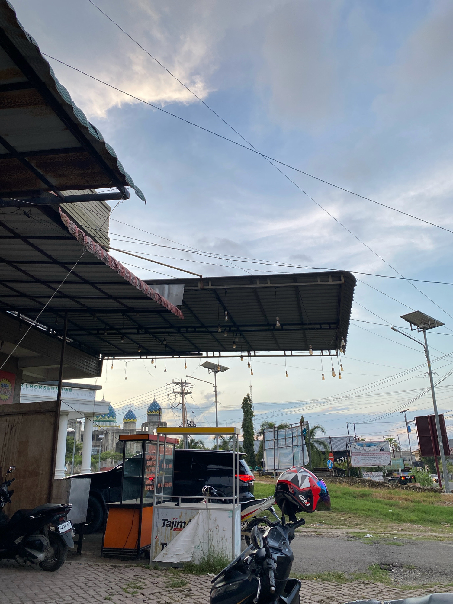 The image depicts a street scene in Lhokseumawe, Indonesia, likely during the late afternoon or early evening given the soft light and cloudy sky. In the foreground, there are two motorcycles parked on a brick-paved area. One motorcycle, a dark scooter, is positioned on the left side of the frame. Another motorcycle, also dark, is on the right, with a red, white, and black helmet resting on its handlebars.
Behind the motorcycles, a small orange food stall with glass display cases is visible. A white tarpaulin covers part of the stall. Further in the background, a black SUV is parked. Dominating the midground and background are a series of structures. To the left, a building with white columns and signage that appears to include "LHOKSEUMAWE KEBANGSAAN" is prominent, with blue-domed structures, possibly part of a mosque, visible behind it. A tall, slender tree stands to the left of this building.
The environment is characterized by overhead wires crisscrossing the sky, a street lamp with a solar panel on the right, and some vegetation and open ground beyond the paved area. The sky is filled with a mixture of blue sky and scattered clouds, with a hint of sunlight breaking through. A large, weathered metal awning extends over the area in front of the building on the left, supported by metal beams and illuminated by strings of bare light bulbs. The overall impression is that of a bustling urban or semi-urban environment, possibly a market or roadside area.