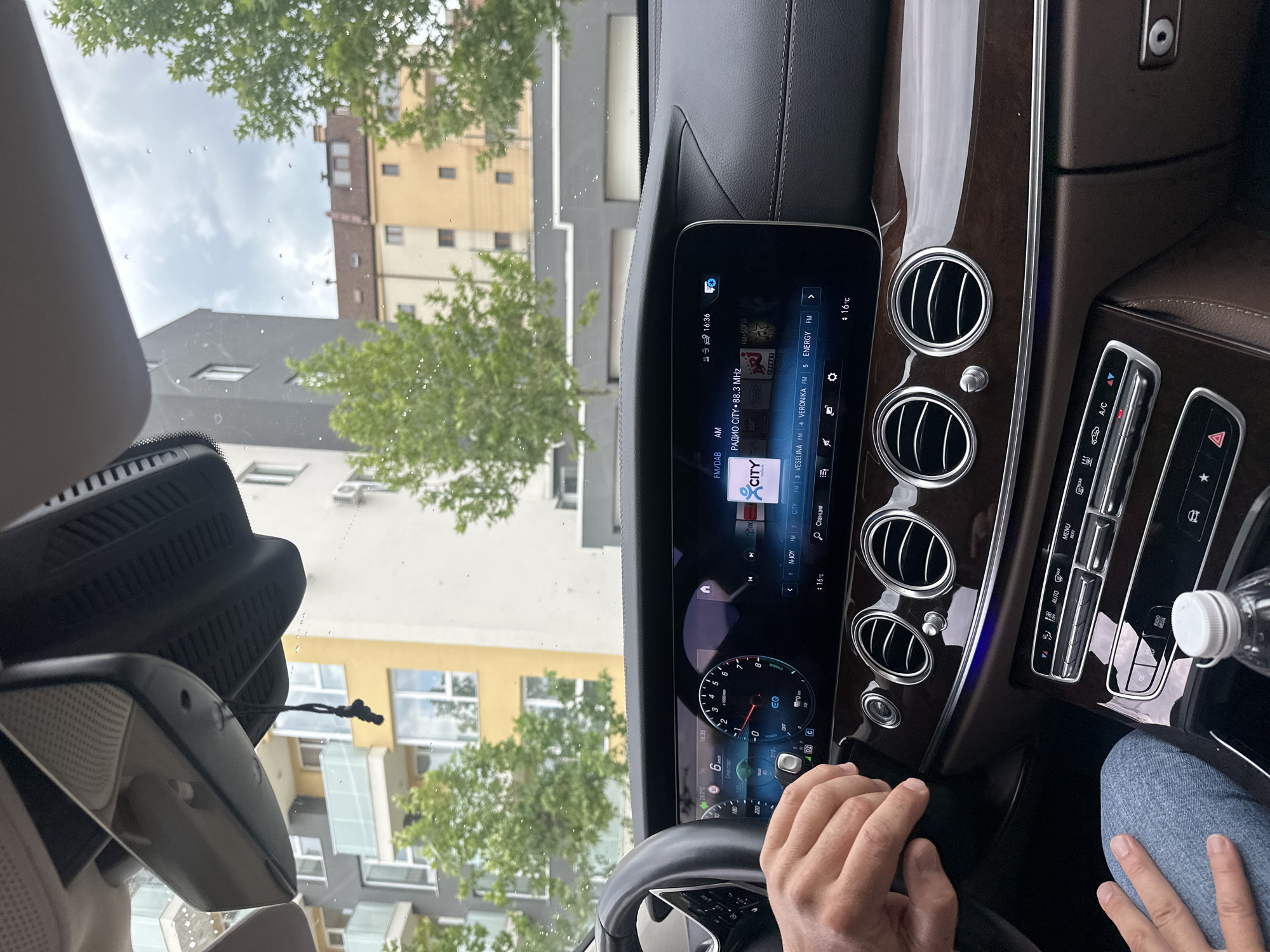 The photo shows the interior of a Mercedes-Benz car, specifically the dashboard and steering wheel, with a driver's hand on the wheel. The car's infotainment screen displays radio information, showing "РАДИО CITY" at 88.3 MHz, along with other radio stations such as "N-JOY", "CITY", "VESELINA", and "ENERGY". The dashboard has air vents and climate control buttons. A water bottle is also visible on the right side of the dashboard. Outside the car, through the windshield, buildings and trees are seen. Based on the radio station name and display language, it is likely in Blagoevgrad, Bulgaria. The car's speedometer shows a reading of 6 km/h. The time displayed on the screen is 16:36.