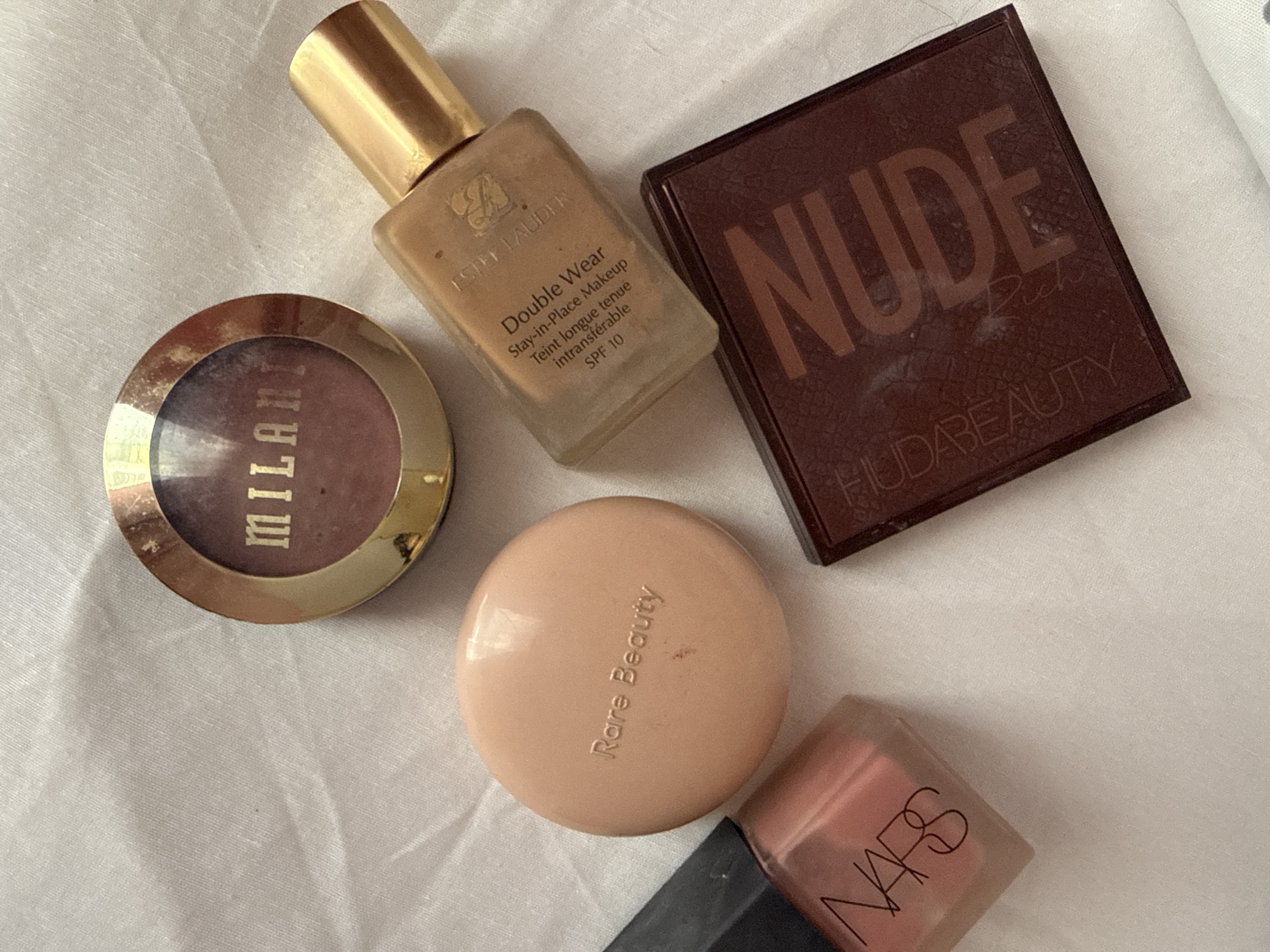 The image shows an arrangement of makeup products on a white surface, likely a cloth. The products include:
* A Milani blush in a gold compact
* A bottle of Estee Lauder Double Wear foundation with a gold cap
* A Huda Beauty Nude eyeshadow palette
* A Rare Beauty product in a round, light beige compact
* A NARS product, possibly a blush or lip product, in a clear square container with a black lid.
The image appears to be taken indoors under bright lighting.