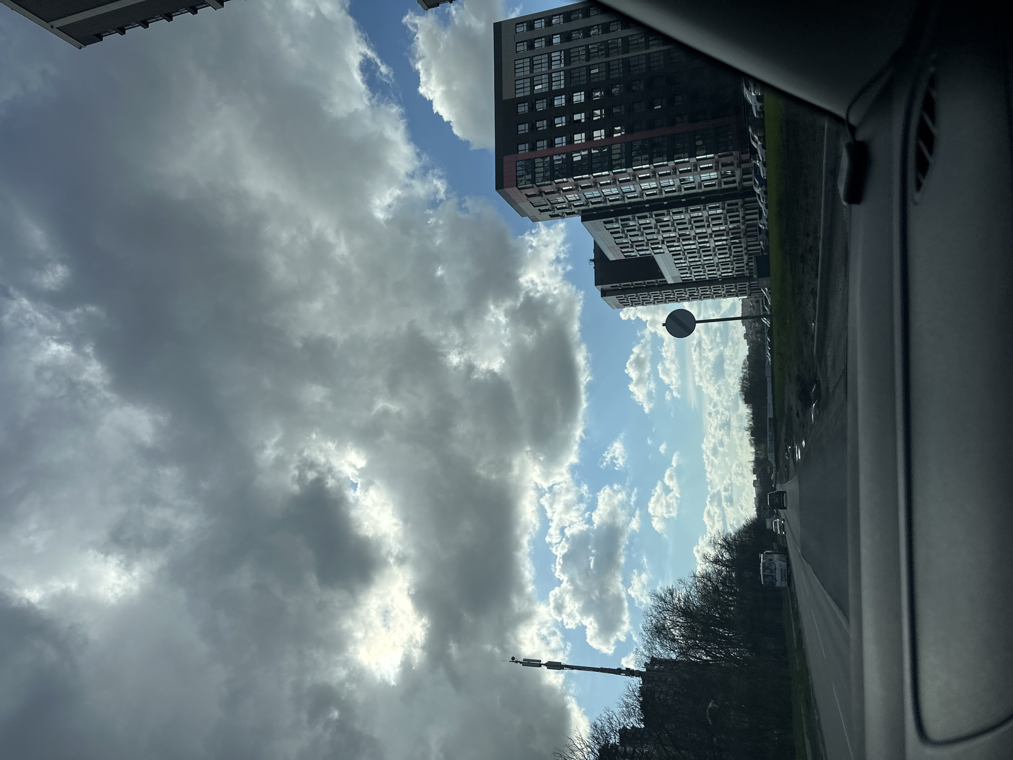 The image is taken from inside a vehicle, likely a car, showing a view of the city Rostov-on-Don, Russia, with a cloudy sky overhead. To the left, large, dark clouds dominate the sky. Between the clouds, patches of blue sky are visible. High-rise apartment buildings are present in the background, indicating a modern urban area. The road stretches into the distance, lined with trees and a few cars are visible in the distance. The interior of the car is visible on the right side of the frame. A streetlight with a circular reflector is observed along the road. A communication tower stands near the treeline.