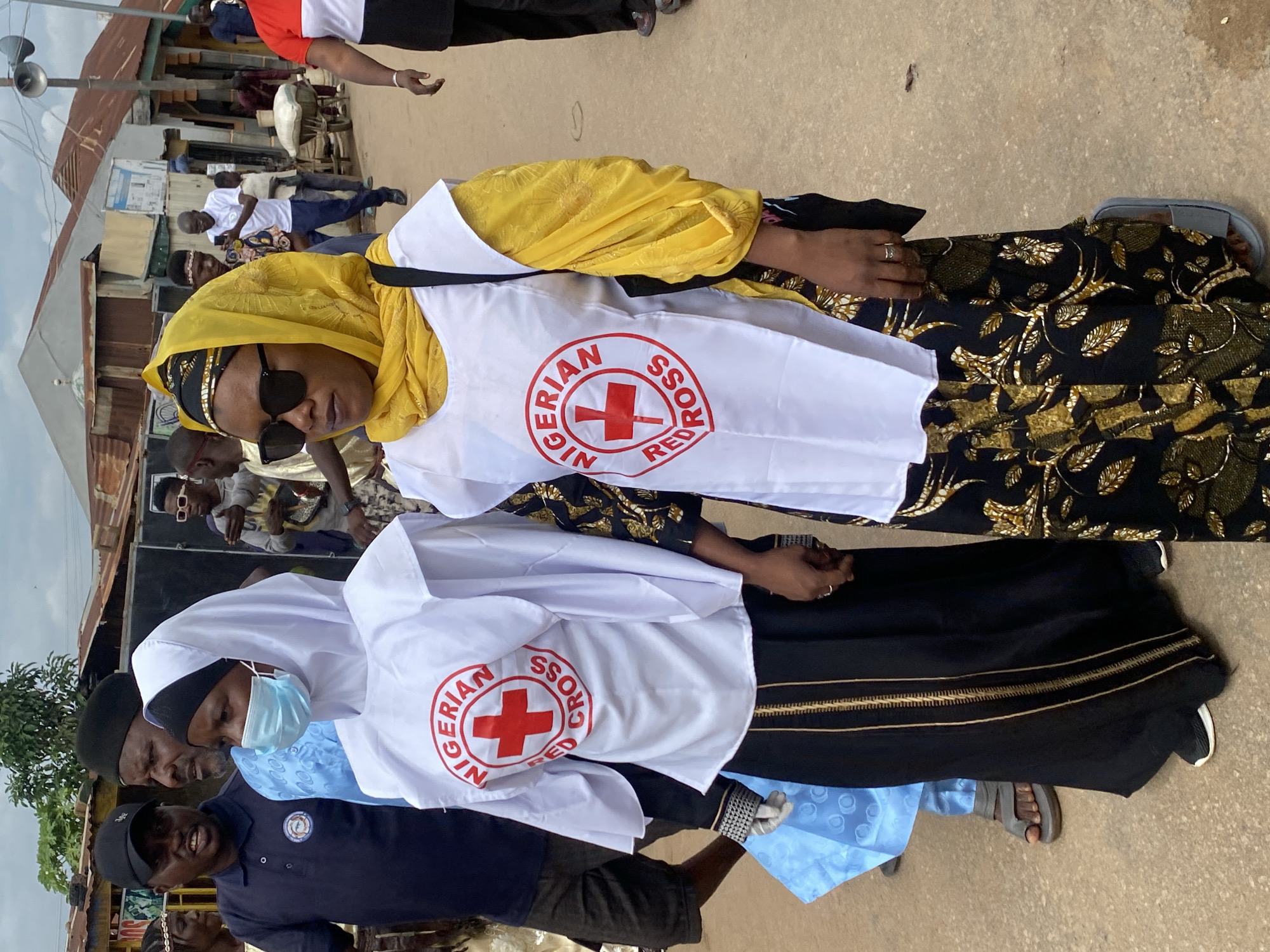 Two women are walking side-by-side outdoors. Both are wearing white vests with the Nigerian Red Cross logo over their clothing. The woman on the left wears a white hijab, a light blue beaded headscarf, and a black skirt with gold trim. She also has a blue face mask on. The woman on the right wears a yellow headscarf and sunglasses. She is wearing a black skirt with a gold leaf pattern. Several other people are visible in the background, including men in traditional clothing and a man in a dark blue polo shirt with a cap. The setting appears to be a street in Rigachikun, Nigeria, with buildings and structures that suggest a market or residential area. The ground is paved with concrete.