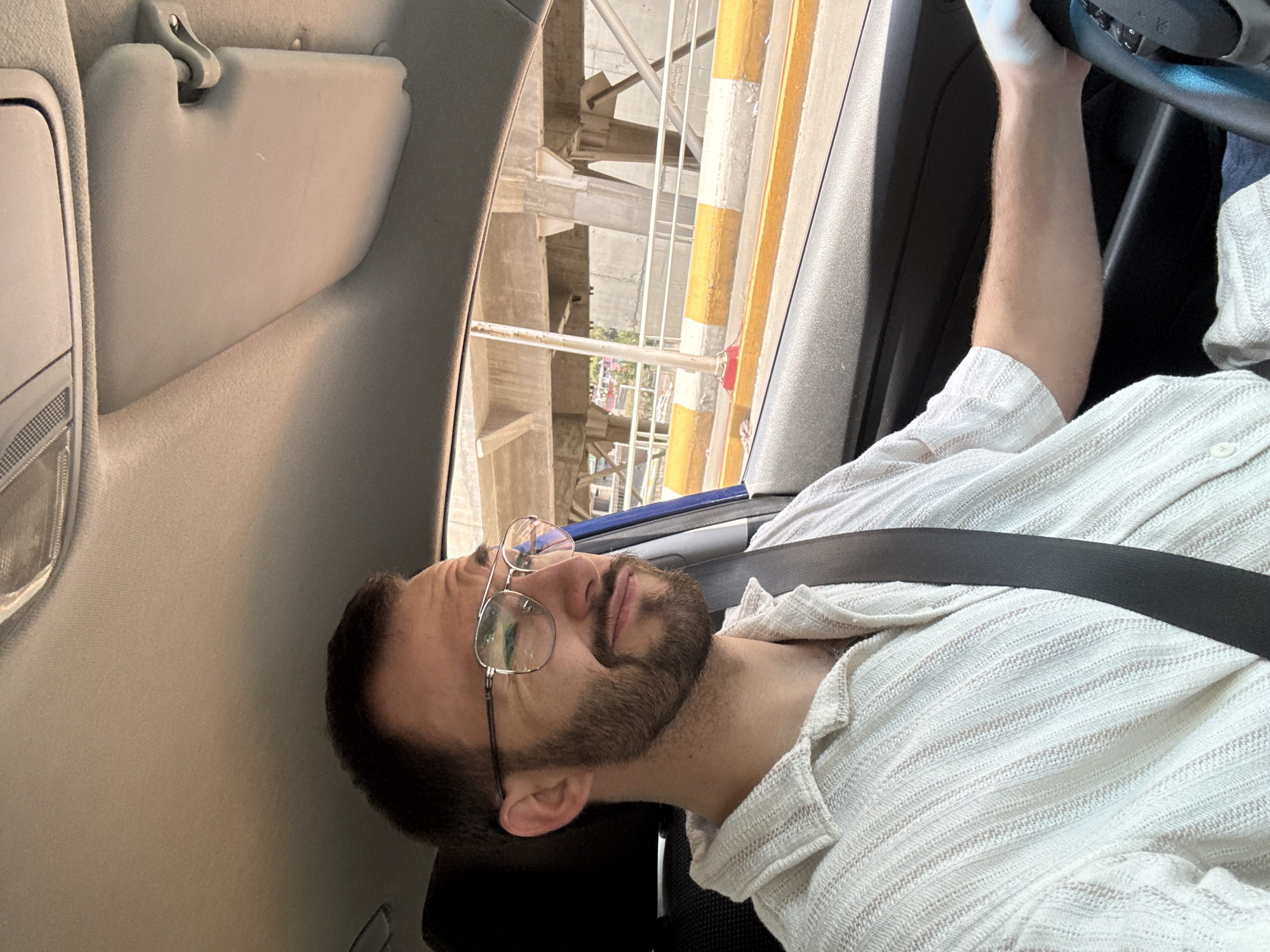 A man with glasses and a beard is sitting in the driver's seat of a car, wearing a seatbelt and holding the steering wheel. The car is driving underneath a highway overpass. The location is likely in Mexico City, Mexico, given the urban environment and the highway infrastructure.