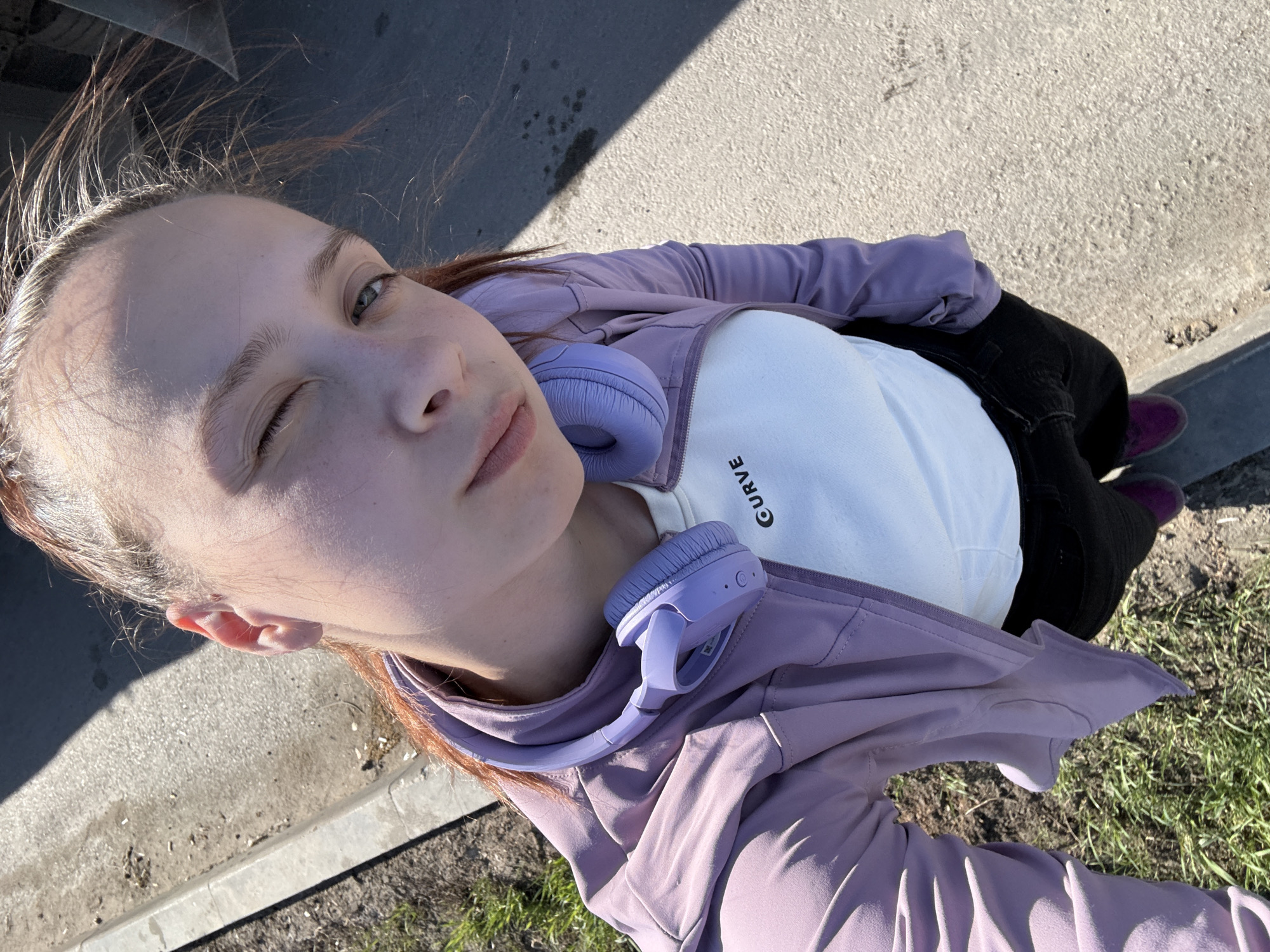 A young person takes a selfie while standing outdoors. The individual has fair skin, one eye winking shut, and is wearing a white t-shirt with the word "CURVE" visible, a lavender jacket, black pants, and purple socks. Headphones of a similar lavender color are draped around their neck. The person is standing on a concrete surface near a patch of grass, likely in an urban setting. The location is possibly in Verkh-Sosnova, Russia.