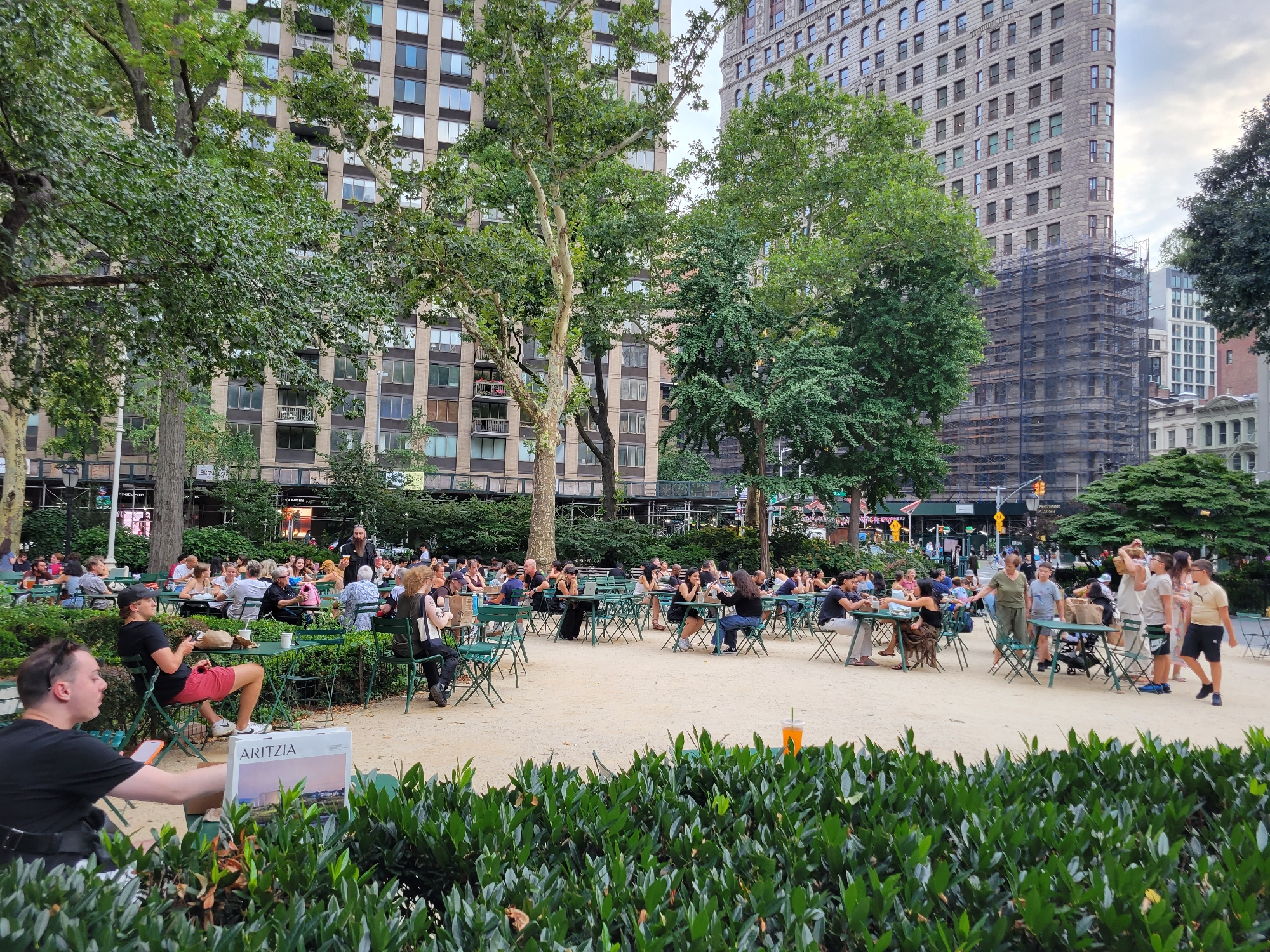 The image depicts a scene in Bryant Park, New York City, United States of America. In the foreground, a person in a black t-shirt is seated, looking to the left, with their legs extended. A magazine with the "ARITZIA" logo is visible on the ground in front of them. A lush green hedge runs across the bottom of the frame.
In the midground, several people are seated at green tables and chairs scattered across a gravel-covered area, suggesting an outdoor dining or gathering space. Some individuals are engaged in conversation, while others appear to be eating or drinking. A variety of trees with green foliage are present, casting shadows on the ground.
In the background, large, multi-story buildings, characteristic of New York City's urban landscape, rise above the trees. One building on the right shows visible scaffolding, indicating construction or renovation. The overall atmosphere appears to be a public park area bustling with people enjoying a daytime outing.