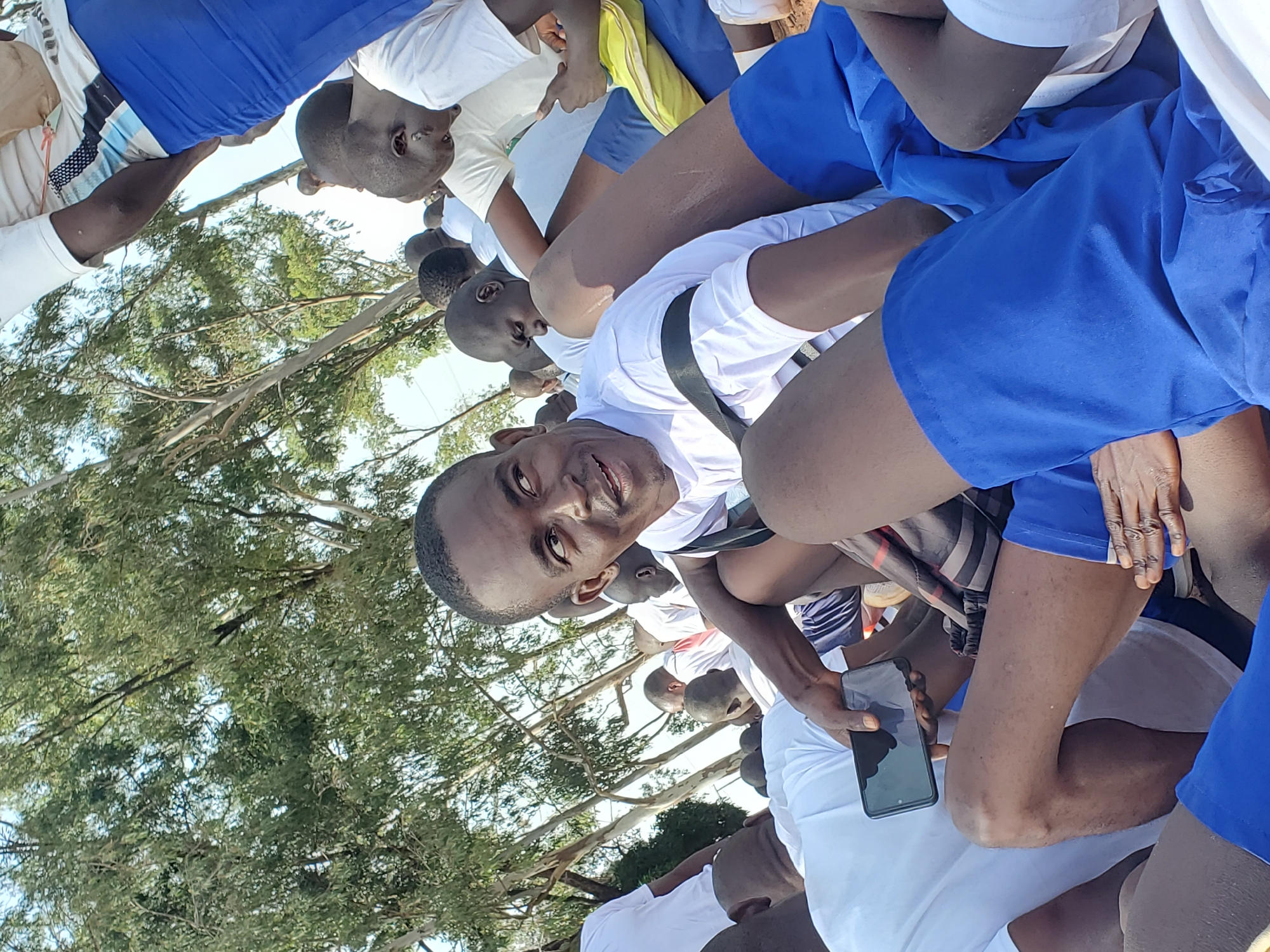 The image shows a group of young men, appearing to be students, gathered outdoors in what seems to be a school or training environment in Barakin Rafin Gora, Nigeria. They are all dressed in similar white t-shirts and blue shorts or trousers. The orientation of the image is sideways.
The individuals are arranged in a cluster, with some seated and others standing. The focus is on a young man in the foreground, who is looking directly at the camera with a slight smile. He is wearing a white t-shirt and black straps, possibly part of a uniform or equipment. He is holding a black smartphone in his right hand.
The setting appears to be outdoors, with trees and foliage visible in the background, suggesting a rural or semi-rural location. The lighting indicates it is daytime, likely with bright sunlight given the clear visibility and shadows. The mood seems relaxed and friendly, with the main subject appearing approachable and engaged.
Some of the other individuals in the group are also visible, with their faces partially obscured or turned away. They share the same attire, suggesting a shared activity or purpose. The overall impression is of a group of young people participating in an outdoor activity, possibly a school event, sports practice, or communal gathering. There is no visible text in the image.