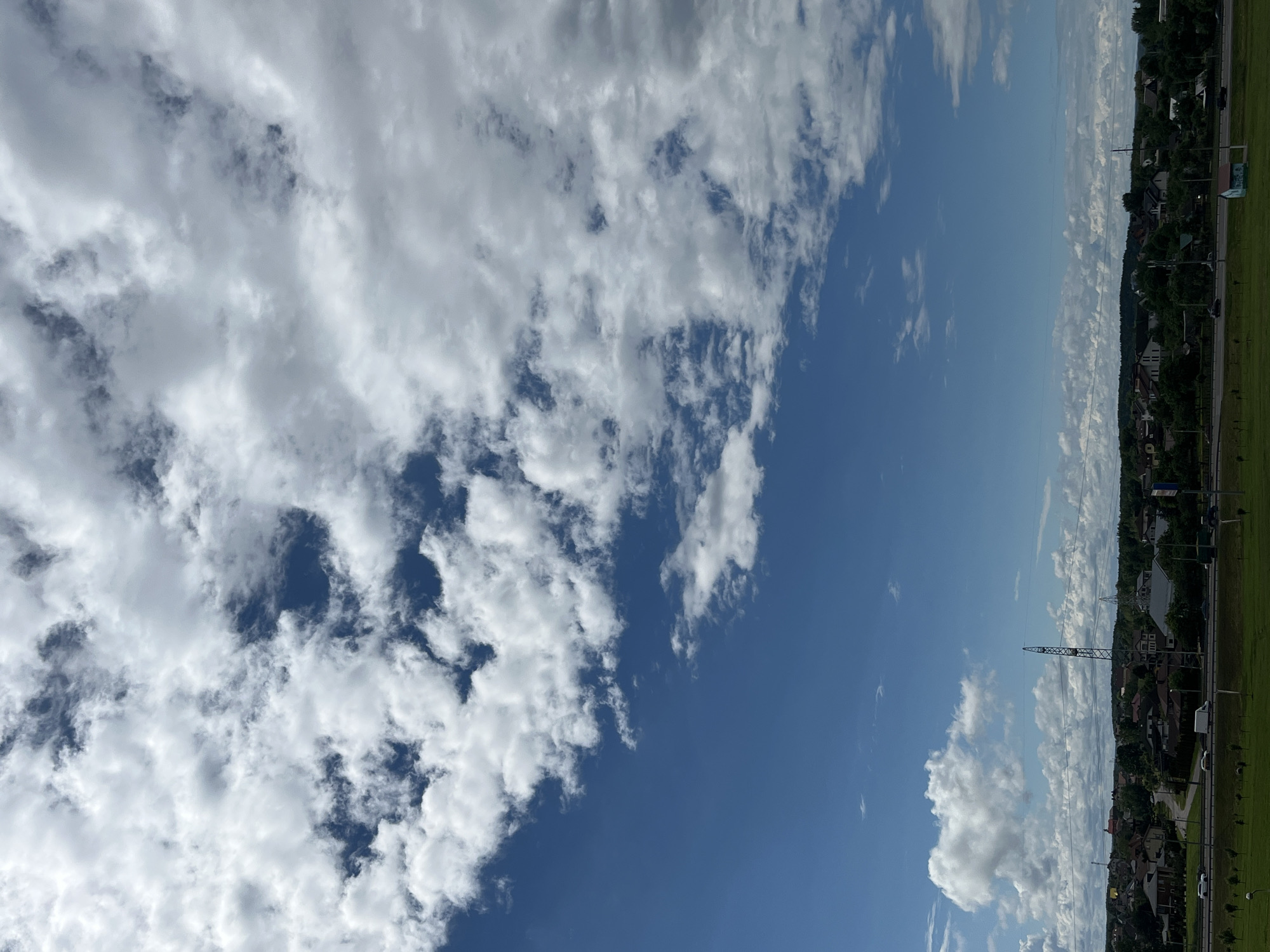 The image captures a suburban landscape under a bright, partly cloudy sky. The upper portion of the frame is dominated by a vast expanse of blue sky interspersed with fluffy white cumulus clouds. Towards the horizon, the clouds become more layered and scattered, creating a textured appearance.
Below the sky, a band of green grass leads to a road with a few vehicles, including a white van and a white car. Beyond the road, a residential area is visible, characterized by a collection of houses with red and brown roofs, interspersed with dense green trees and foliage. A prominent metal transmission tower stands tall among the buildings and trees. In the distance, a few more buildings and structures can be seen, suggesting a moderately developed area. The overall impression is that of a typical suburban setting on a clear day.
